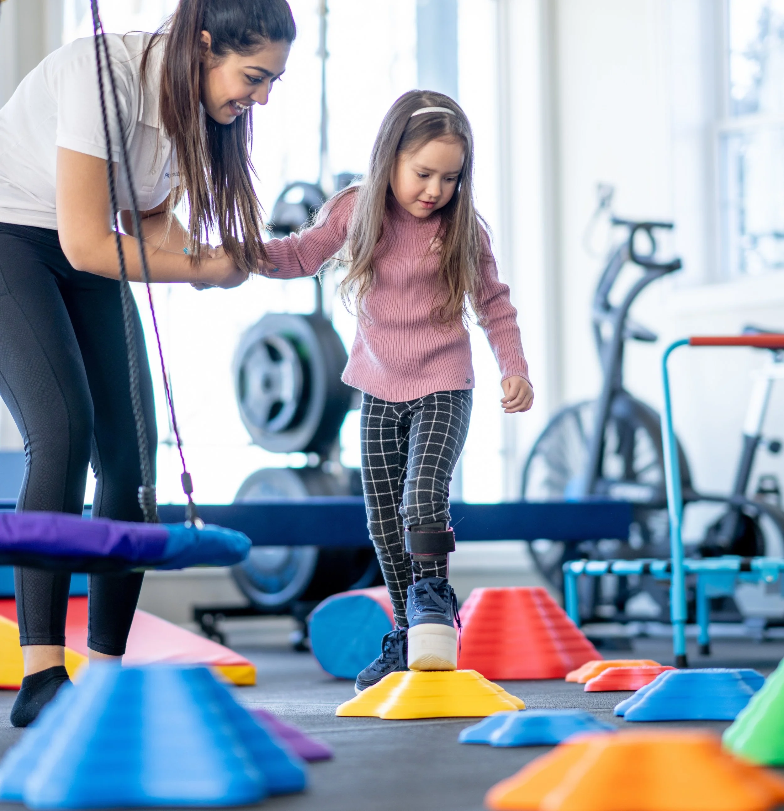 A young girl and a woman participating in an indoor activity, stepping over colorful cones set up on the floor during a fitness or physical education class.