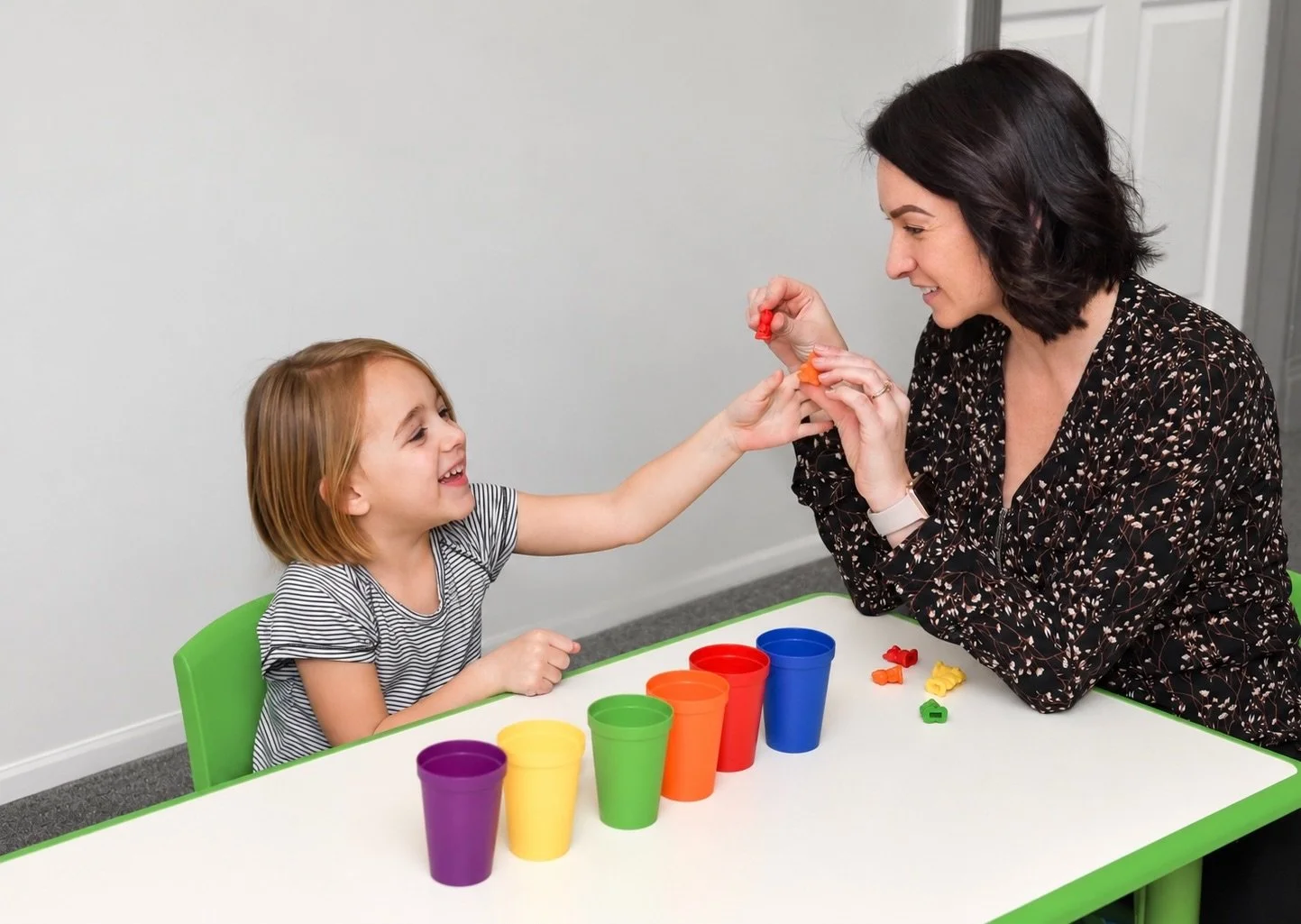 A woman and a young girl sit at a table with colorful cups. The woman is holding small red and orange objects, offering them to the girl who is smiling and reaching out to take them.