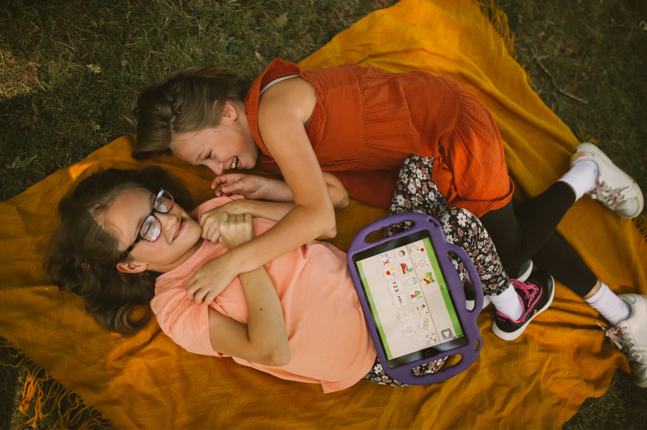 Two young girls lying on a yellow blanket on the grass, interacting with a tablet device. One girl has glasses and dark hair, while the other has light brown hair and is wearing an orange dress.