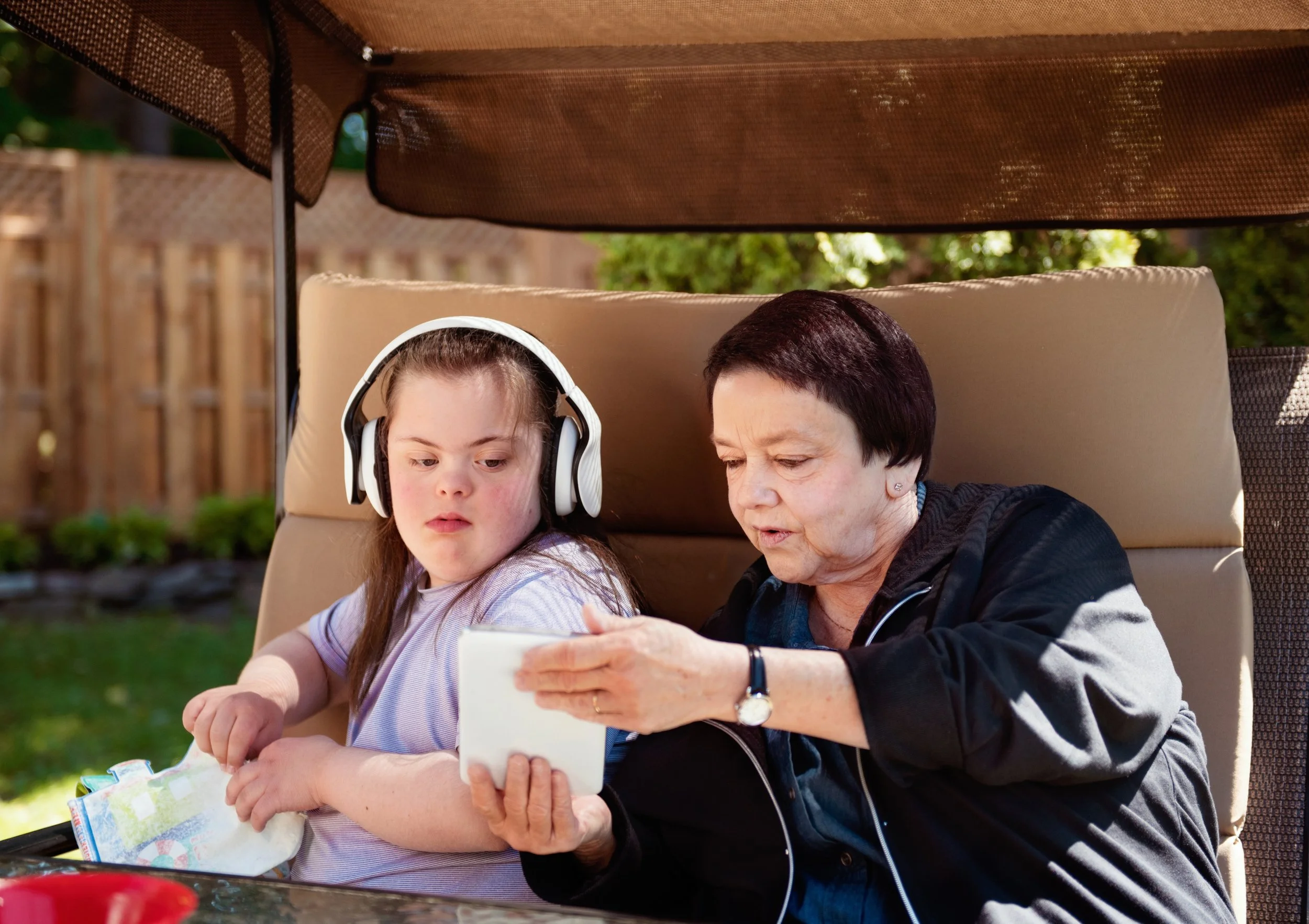 A woman and a girl sitting on a patio with a beige cushion behind them, looking at a tablet device. The girl is wearing headphones and a striped shirt, the woman has short dark hair and is wearing a black jacket. There is a wooden fence and greenery in the background.