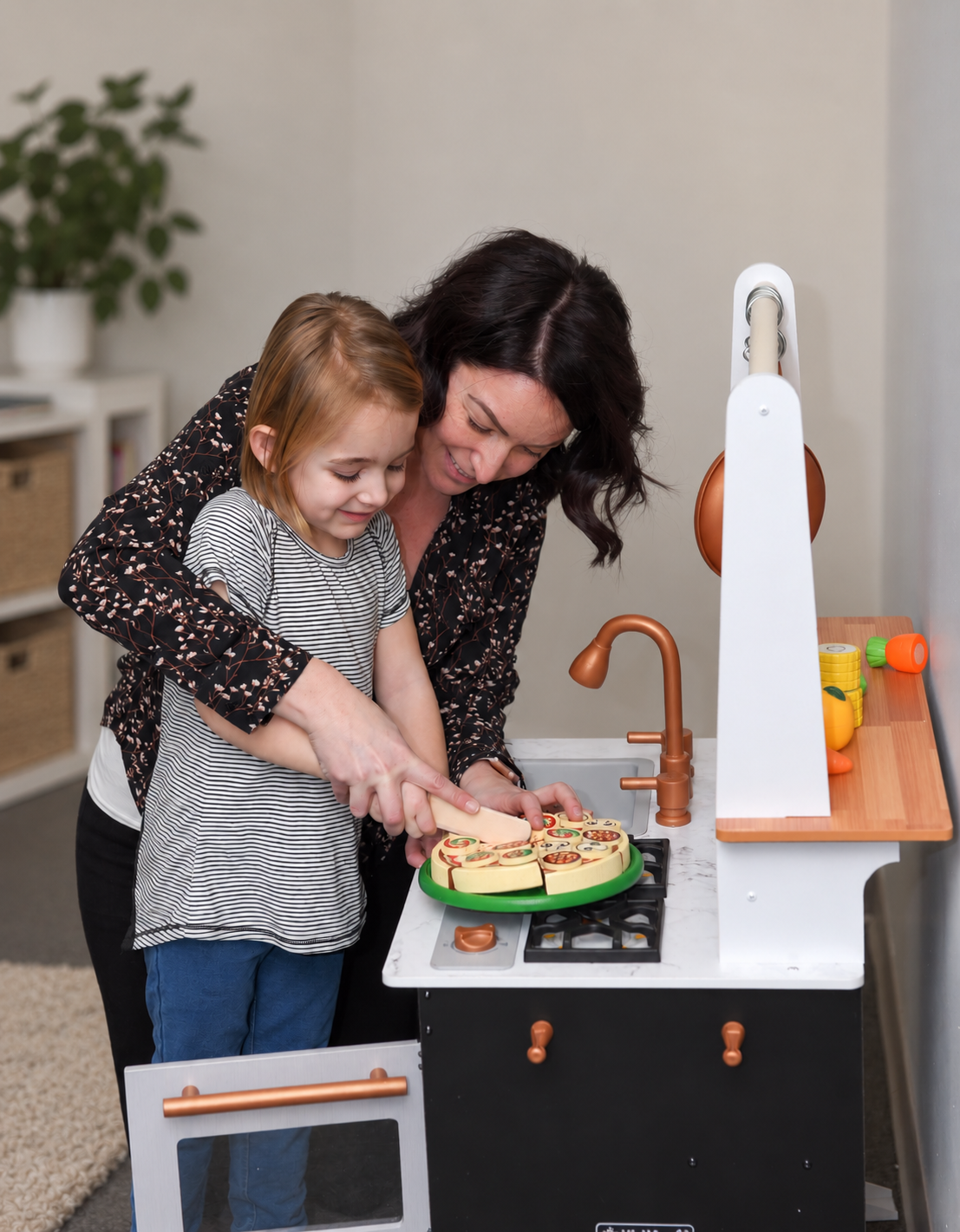 A woman and a young girl playing with a toy kitchen set, pretending to cook together. The woman is helping the girl slice toy food on a green plate.