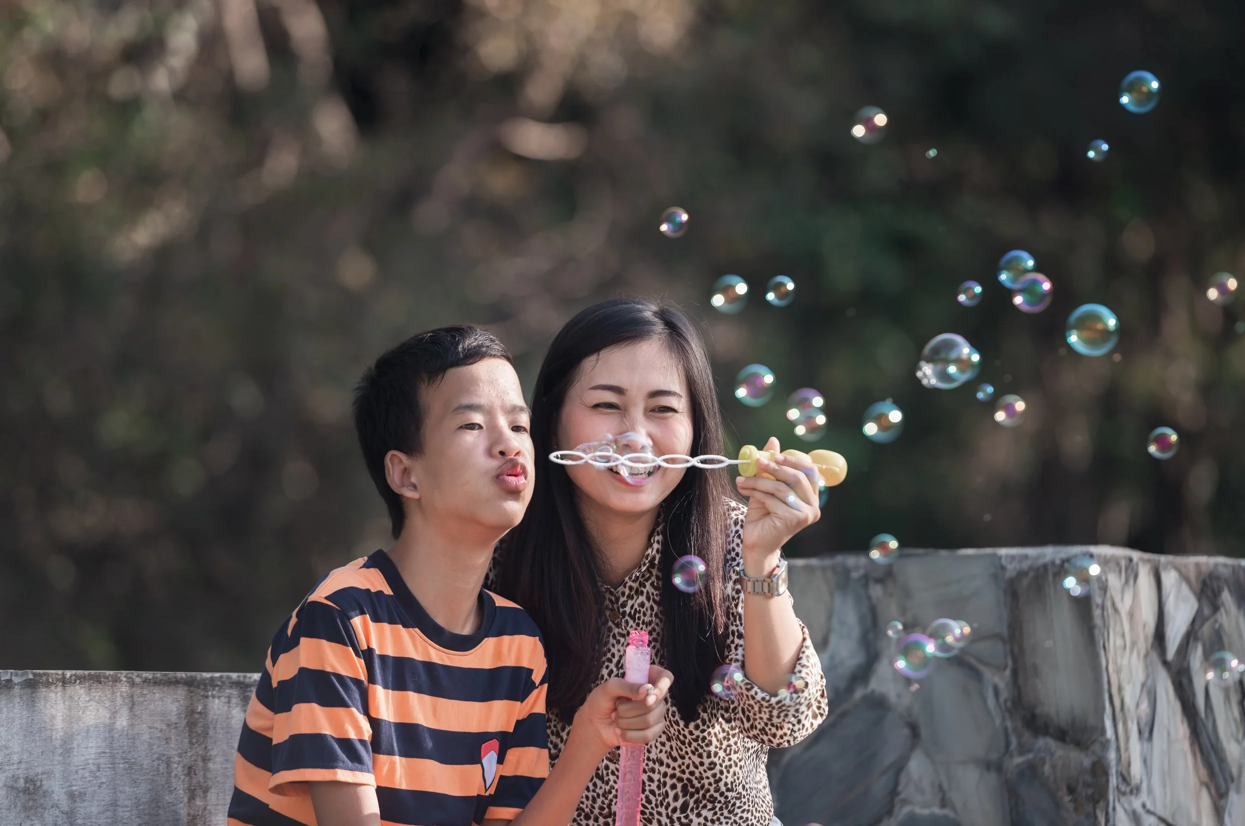 Two people, a woman and a young boy, are outdoors blowing soap bubbles, with the woman smiling and the boy focusing on the bubbles.