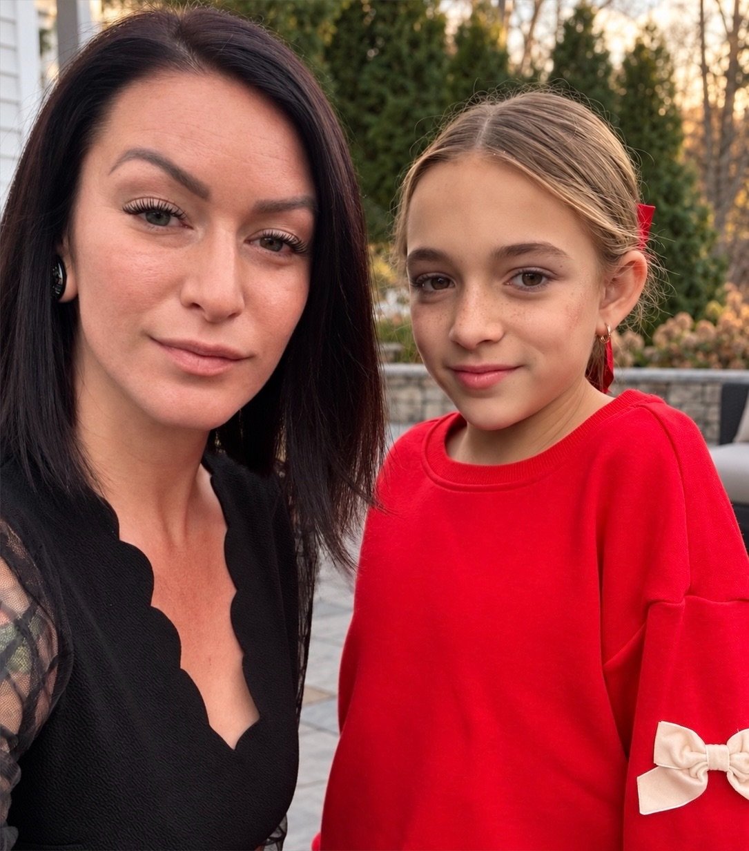 A woman and a girl taking a selfie outdoors during sunset. The woman has dark hair and is wearing a black top with lace sleeves. The girl has blonde hair tied with a red ribbon and is wearing a red sweatshirt with a bow on the sleeve.