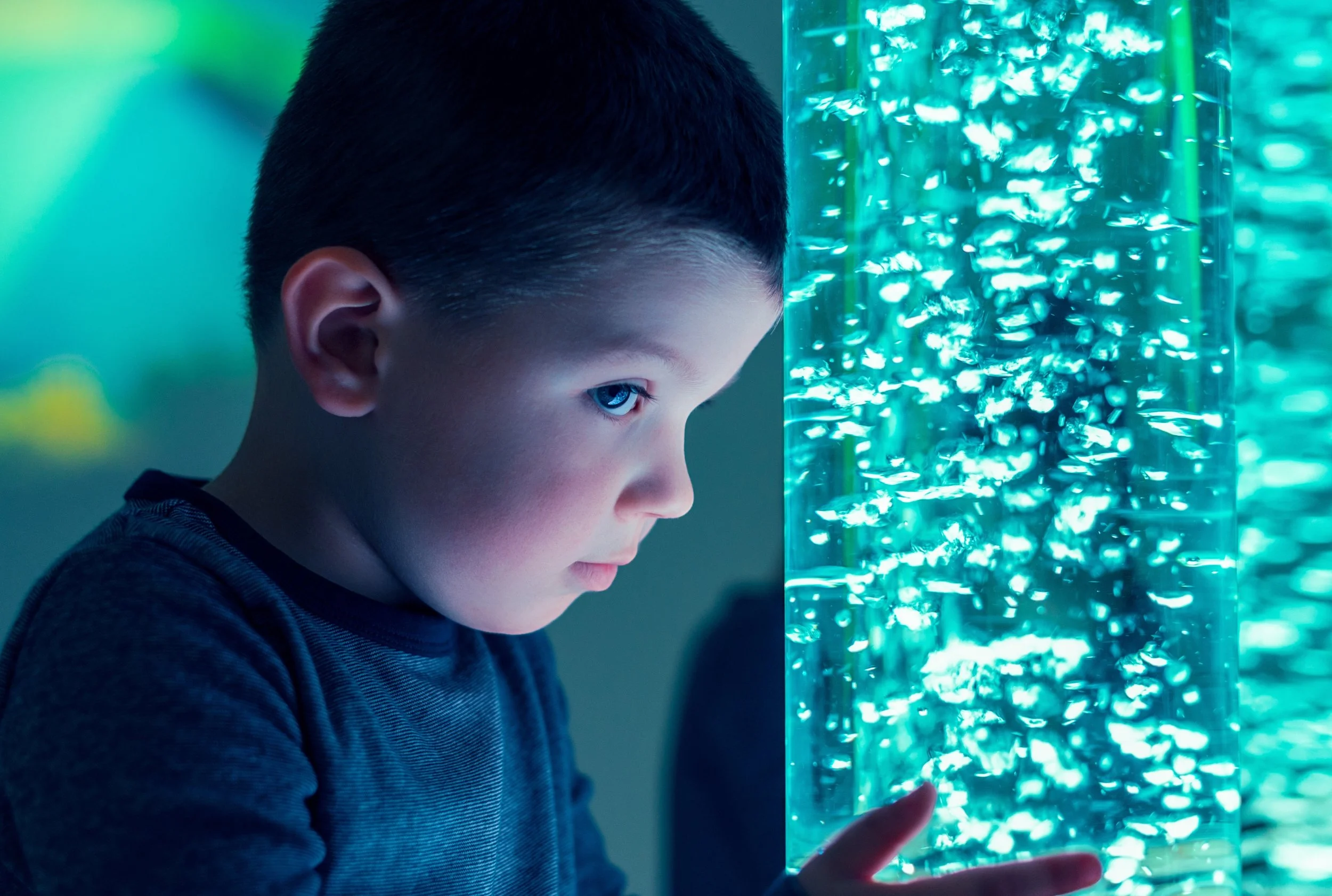 A young boy with short dark hair and light skin, gazing at an illuminated aquarium tank filled with small fish, with his finger touching the glass.