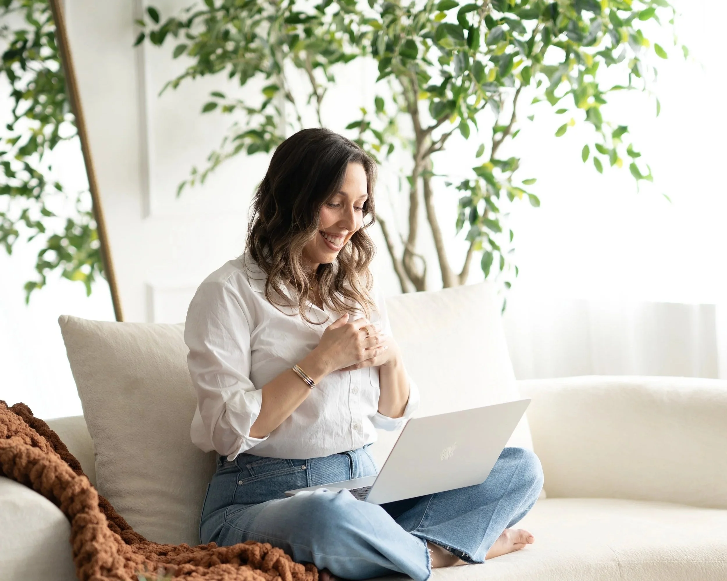 Leslie Gil, LMHC, smiling while using her laptop on a sofa at The DBT Collective, offering DBT therapy, clinical supervision, and professional training in Miami, Florida.