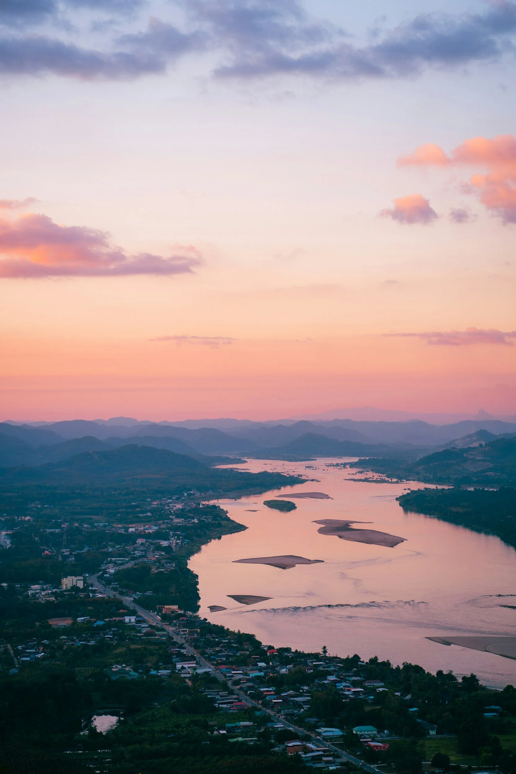 A scenic view of a river winding through a valley during sunset with pastel pink, purple, and orange clouds in the sky, mountains in the distance, and a small town along the riverbanks.