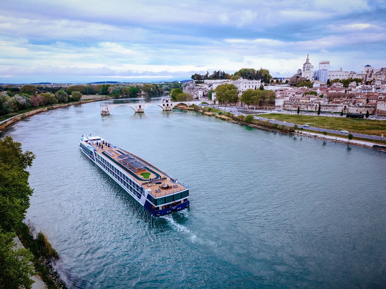 A large cruise boat sailing on the river in a city with historic buildings, bridges, and lush trees, under a cloudy sky.