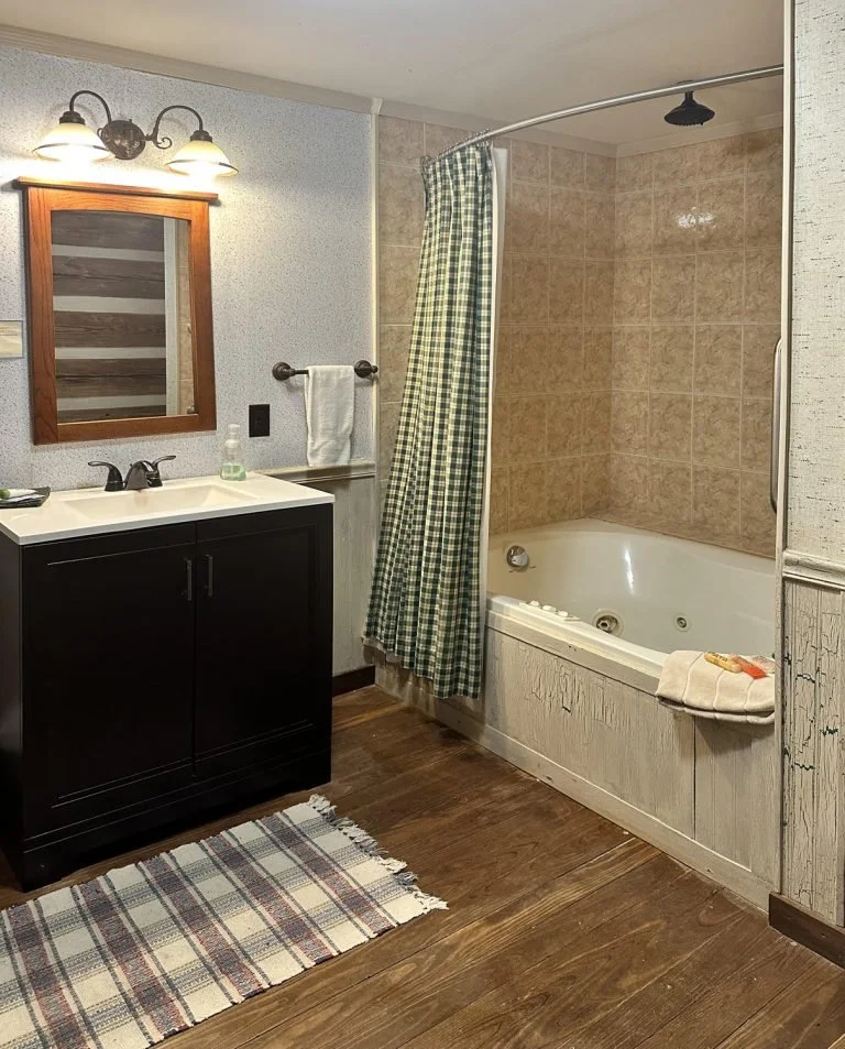 A rustic bathroom with a black vanity, white sink, mirror, wall-mounted light, beige tile shower with a curved rod, green checked shower curtain, whirlpool tub with a towel on the edge, wooden floor, and plaid rug.