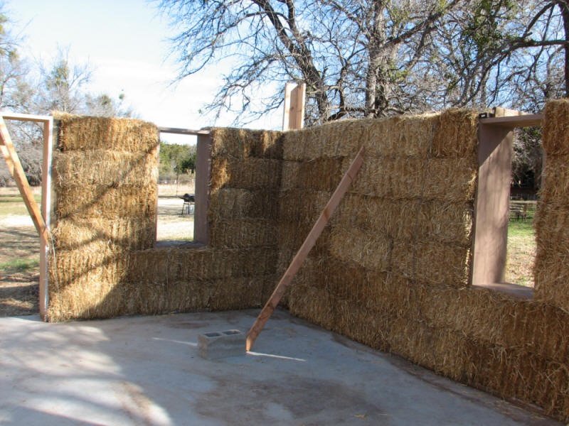 Barn wall under construction with hay bales stacked to form walls, window openings, and support beams, outside on concrete foundation with trees in the background.