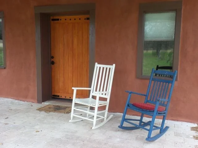 Two rocking chairs, one white and one blue with a red cushion, are placed outside in front of a house with a stucco wall and a wooden door.