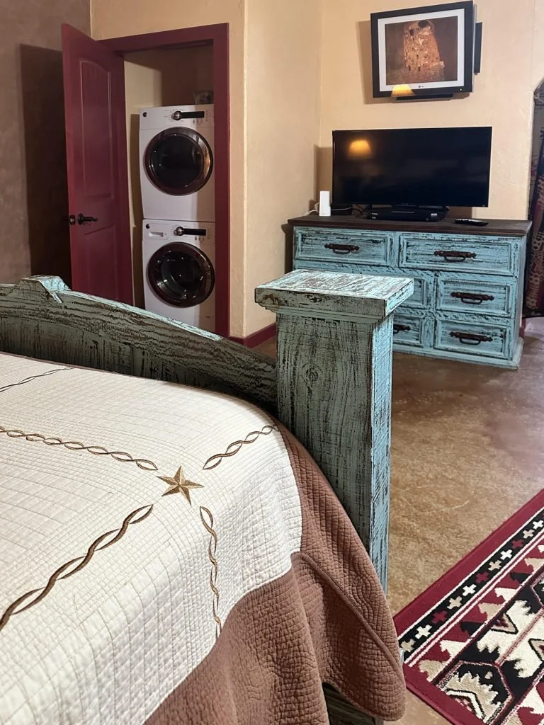 A bedroom with a wooden bed frame, a quilted bedspread, a distressed blue dresser with a flat-screen TV on top, a wall-mounted TV above it, a laundry closet with stacked washer and dryer, a framed picture on the wall, and a red decorative rug on the floor.