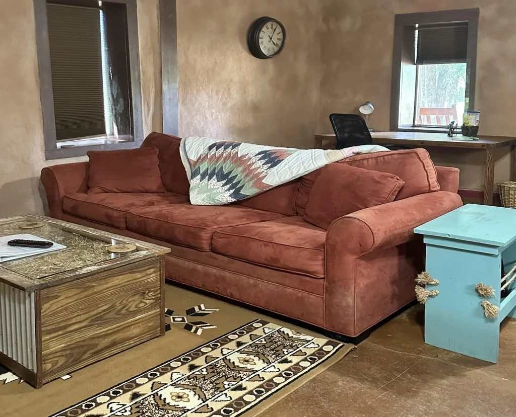 Living room with a salmon-colored sofa, a quilt draped over it, and a wooden coffee table with a remote and notepad on it. There are two windows with open blinds, a wall clock, a desk with a lamp and plants, and a blue side table with decorative rope accents.