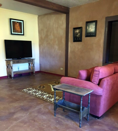 Living room with a pink sofa, a small wooden side table, a television on a decorative stand, framed pictures on the wall, and hardwood flooring.