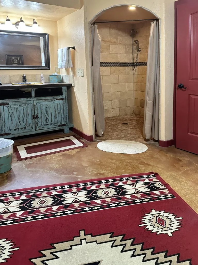 View of a bathroom with a shower, vanity, mirror, and various rugs on the floor, including a colorful southwestern patterned rug.