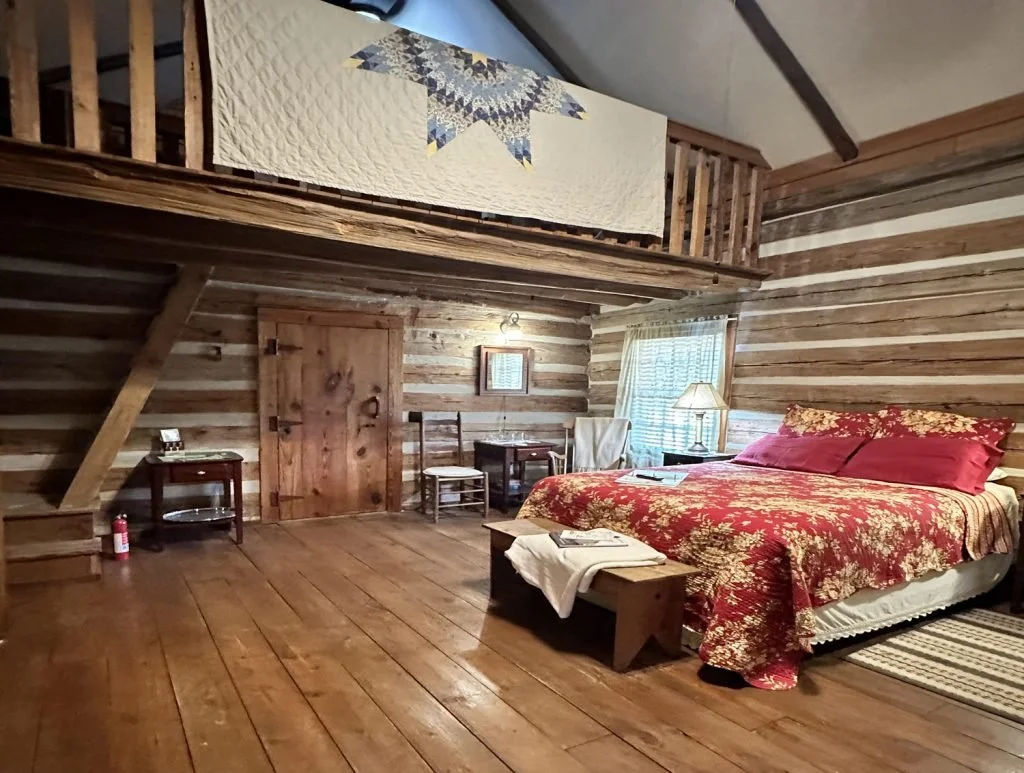 A rustic bedroom with wood-paneled walls and floor, a bed with red and gold floral bedding, a window with sheer curtains, a bedside lamp, a bench at the foot of the bed, and a small wooden door on the wall. There is also a quilt hanging from a loft.