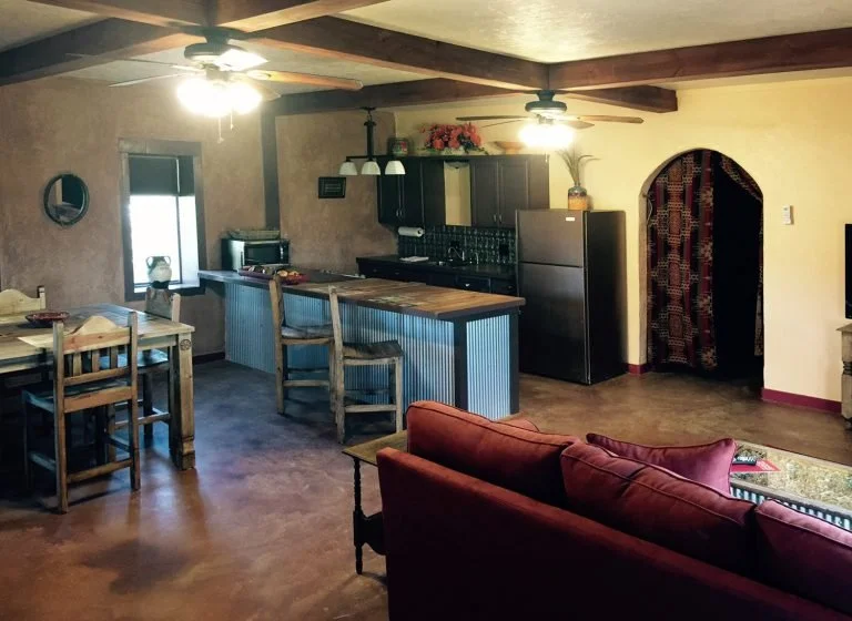 Open-concept living room and kitchen with wooden beams, ceiling fans, a dining table with chairs, a kitchen island with barstools, black cabinets, stainless steel appliances, and a doorway with a patterned curtain.