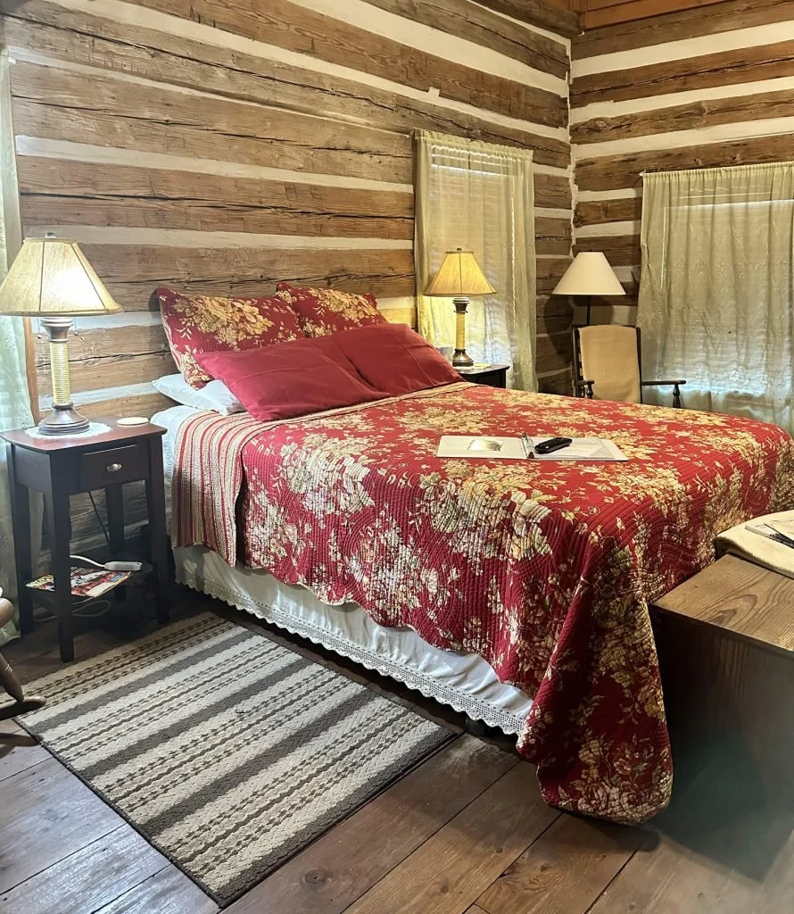 A bedroom with wooden log walls, a bed with a floral red and gold bedspread, matching pillows, and multiple lamps on bedside tables.