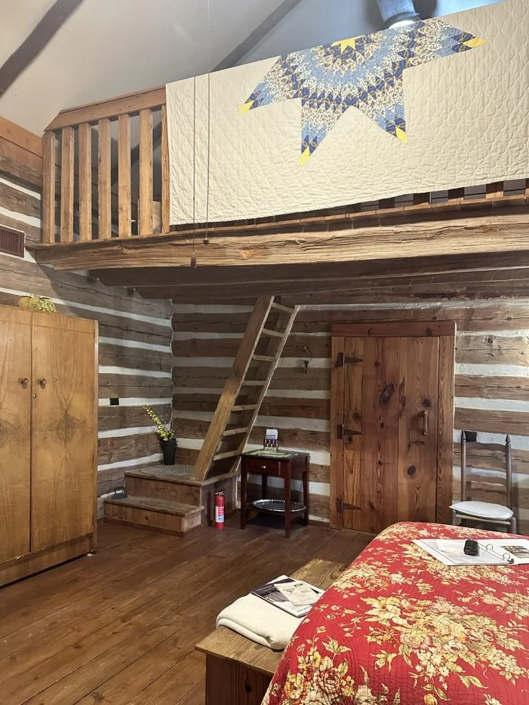 A cozy rustic room with wooden walls and floors, a bed with a red floral quilt, a wooden wardrobe, and a small bedside table. There is a loft area with a quilt featuring a star quilt pattern, accessed by a wooden ladder.