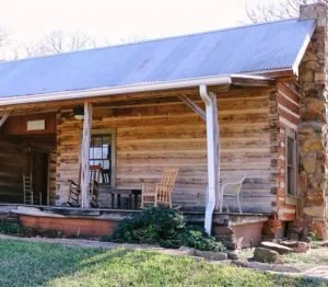 Wooden cabin with a metal roof and small porch, surrounded by grass and trees.