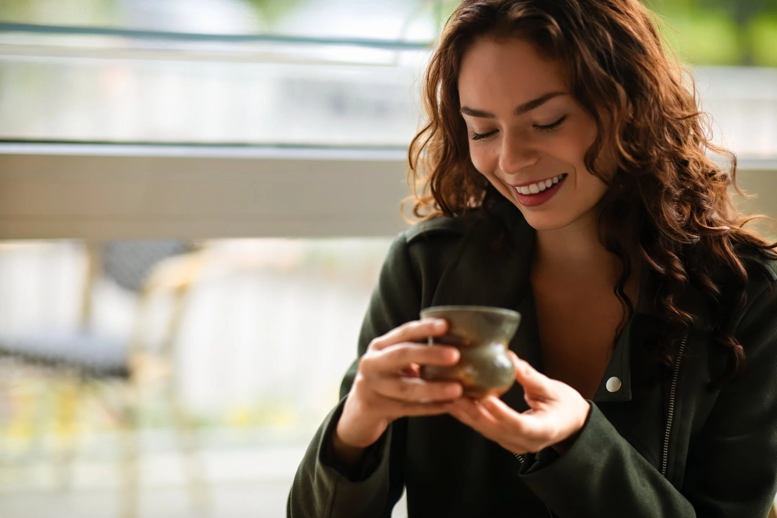 A woman with curly red hair smiling and holding a ceramic cup, sitting near a window with blurred green trees outside.
