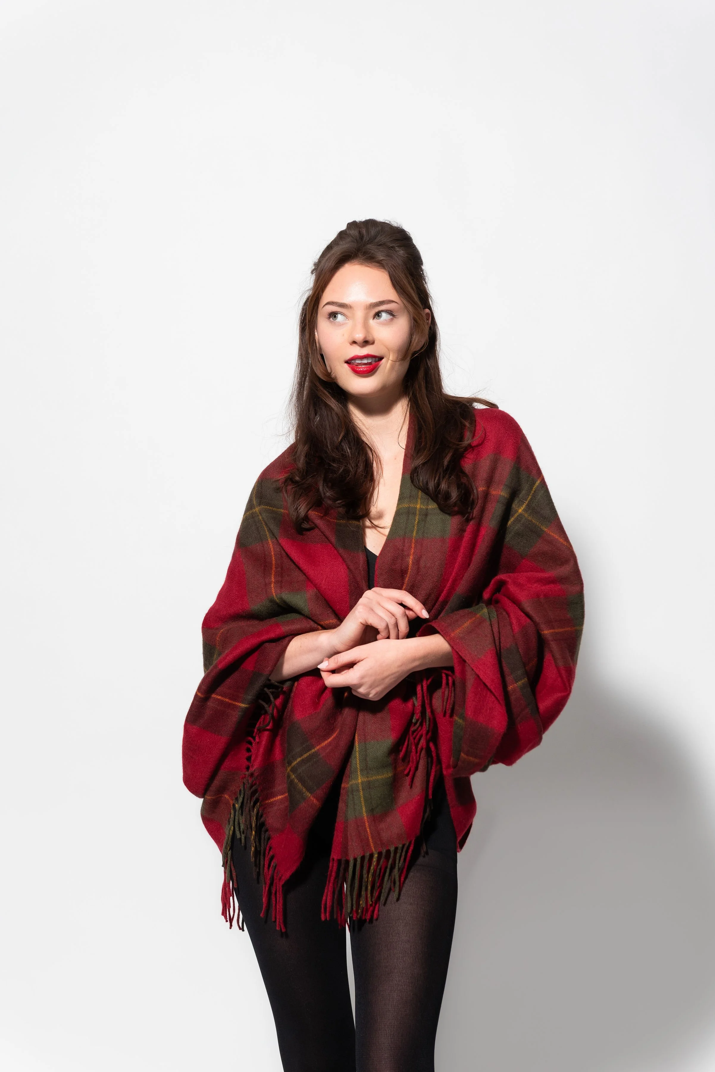 A young woman with long brown hair and red lipstick wearing a red and green plaid shawl, black leggings, and standing against a plain white background.