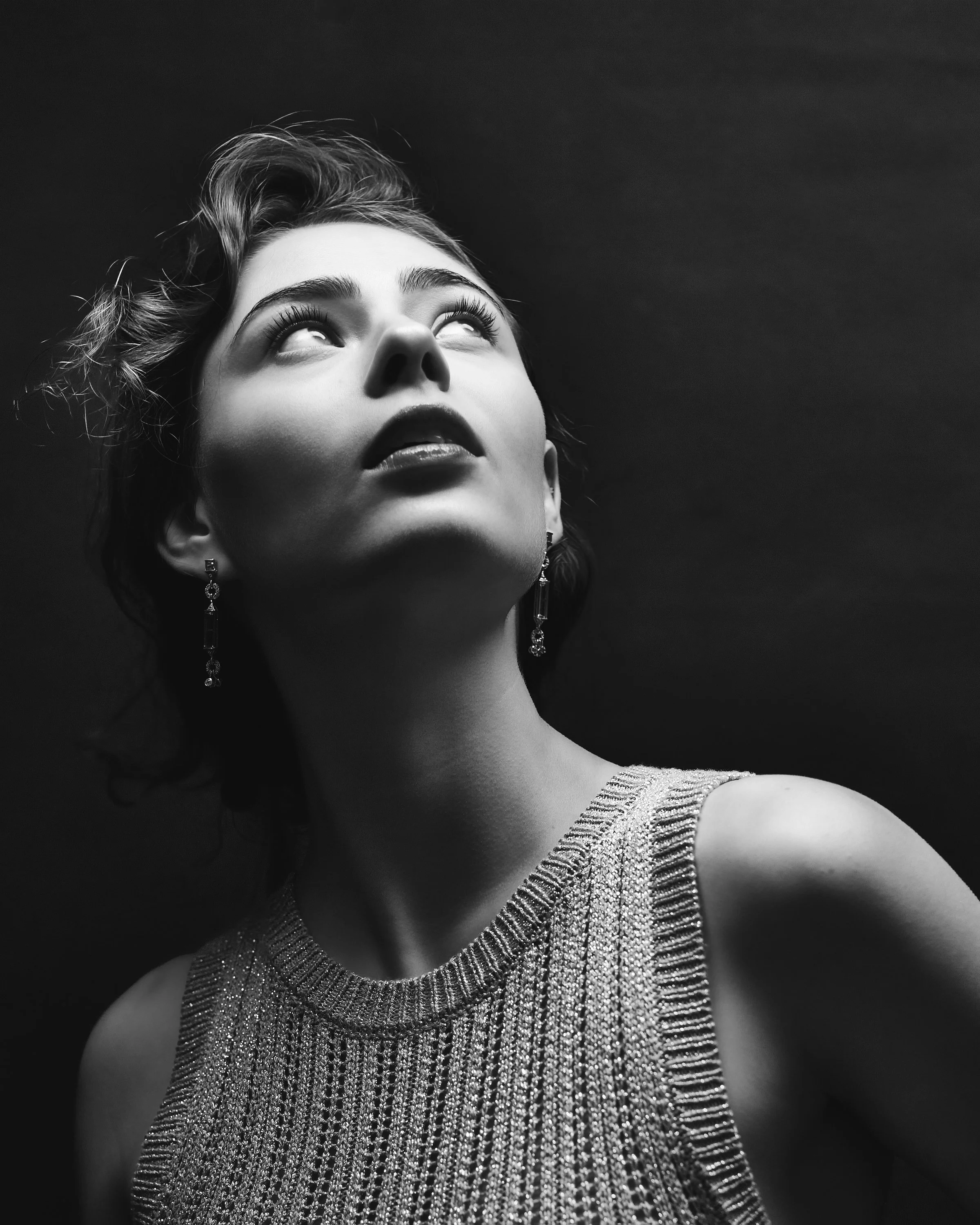 Black and white portrait of a woman with short, styled hair, wearing dramatic makeup, and a shimmery dress with elaborate jewelry, looking upwards with a serious expression.