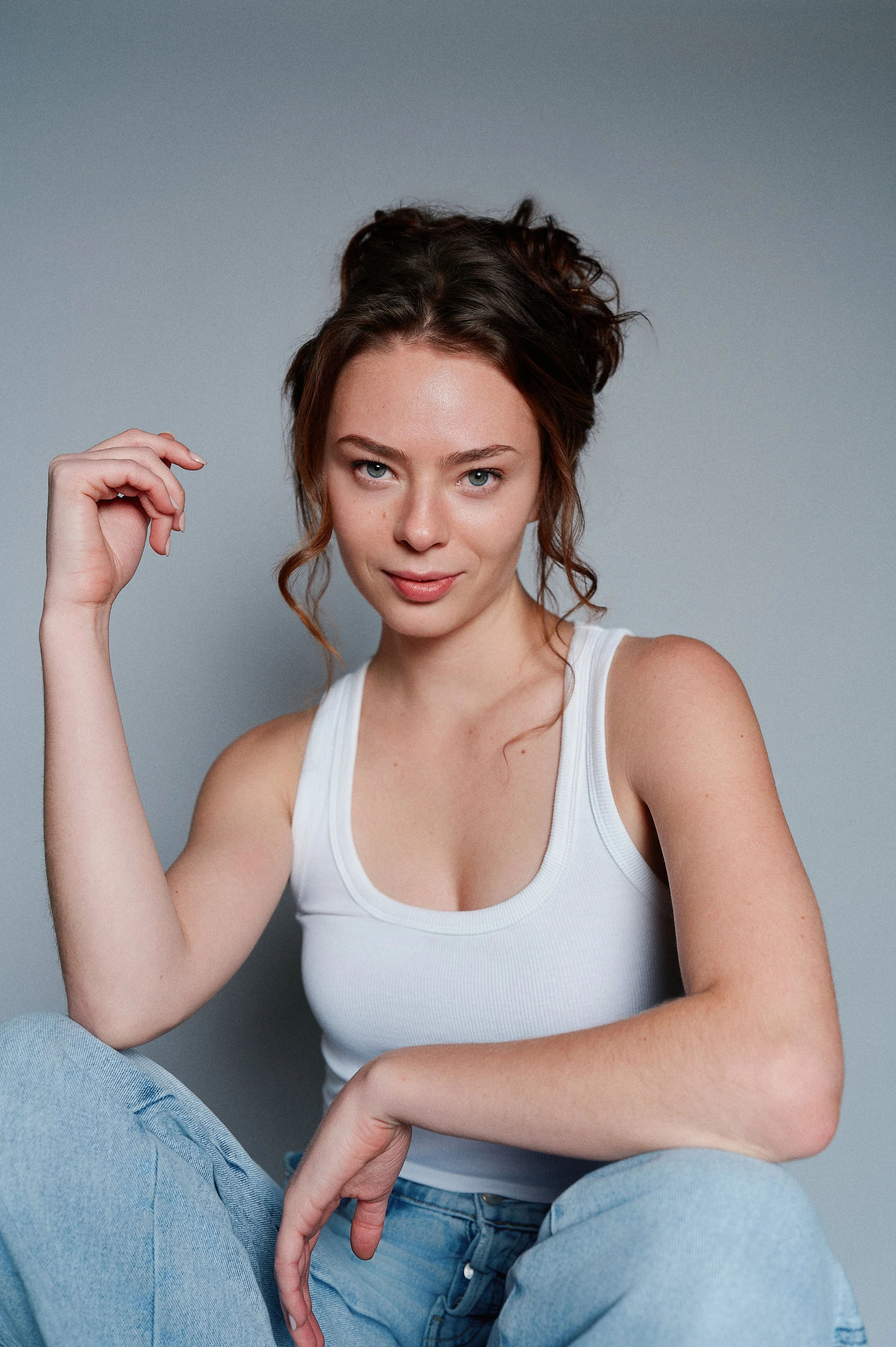 A woman with curly brown hair wearing a white tank top and light blue jeans sitting against a gray background, looking into the camera with her left hand raised near her face and her right arm resting on her leg.