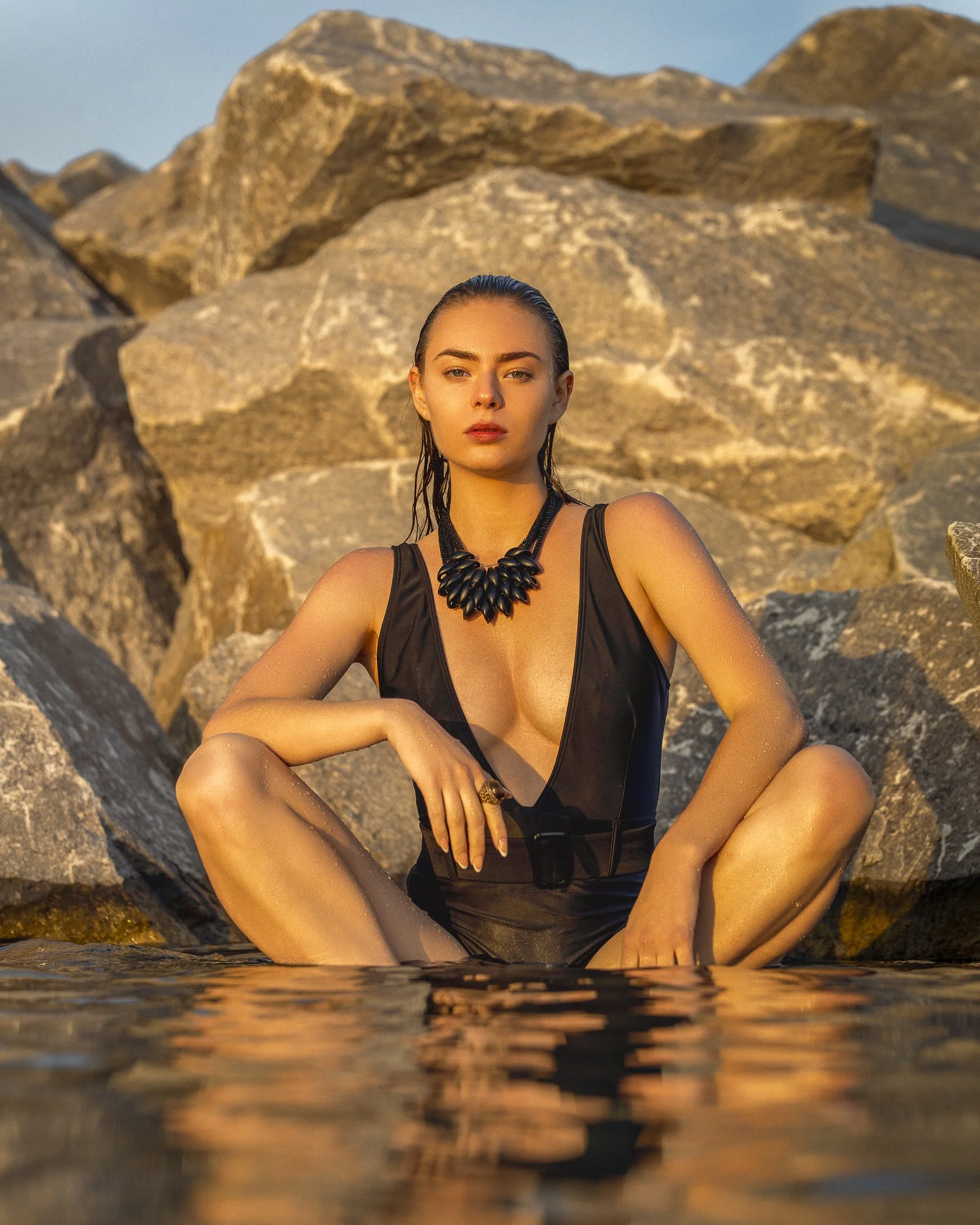 A woman with wet hair and a black swimsuit sits among large rocks at sunset, partially submerged in water.