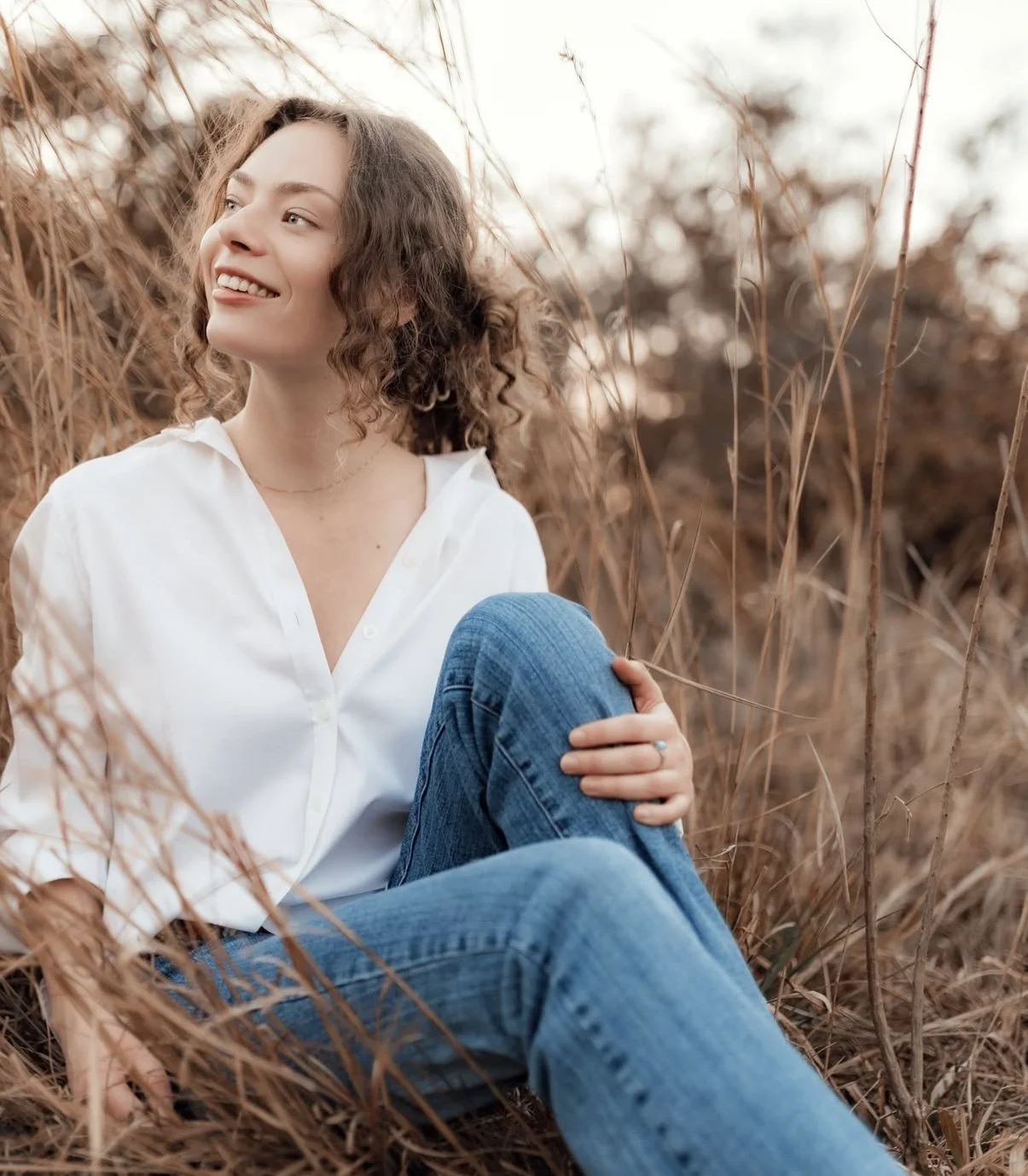 A woman with curly hair sitting among dry grass, smiling and looking to the side, wearing a white shirt and jeans.
