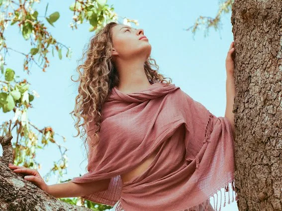 A woman with curly hair leaning against a tree, looking up at the sky, wearing a pink shawl, outdoors with trees and leaves in the background.