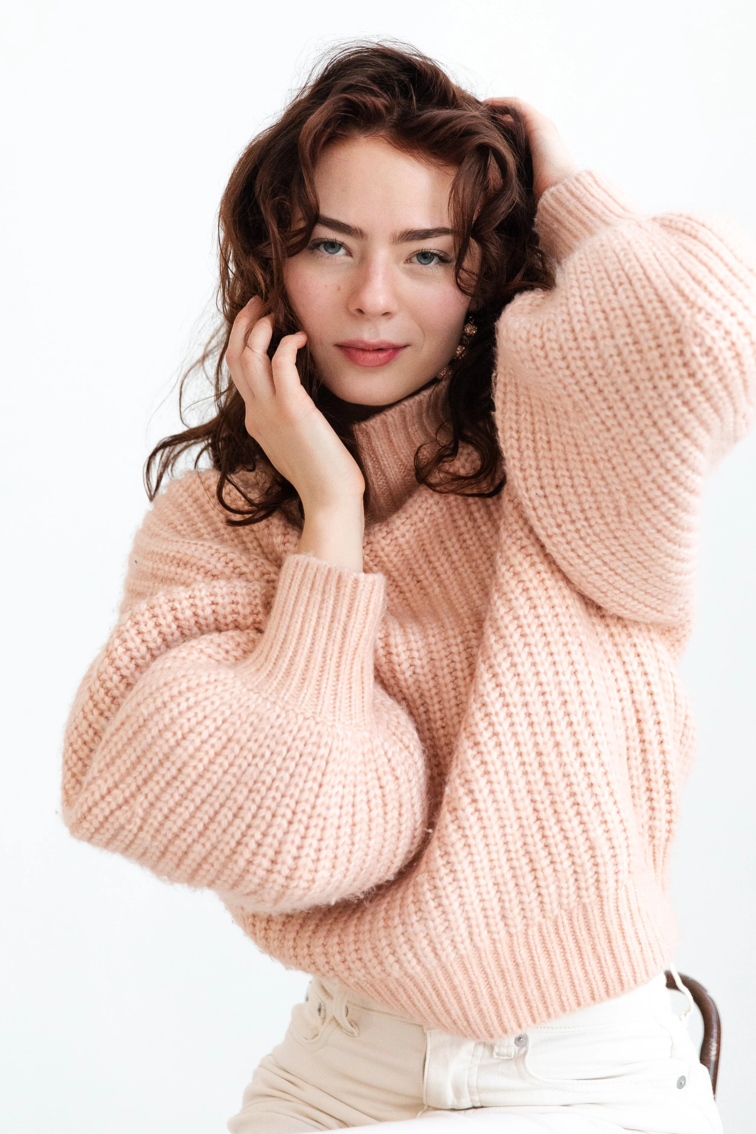 A young woman with curly brown hair and blue eyes, wearing a pink knitted sweater and beige pants, sitting against a white background.