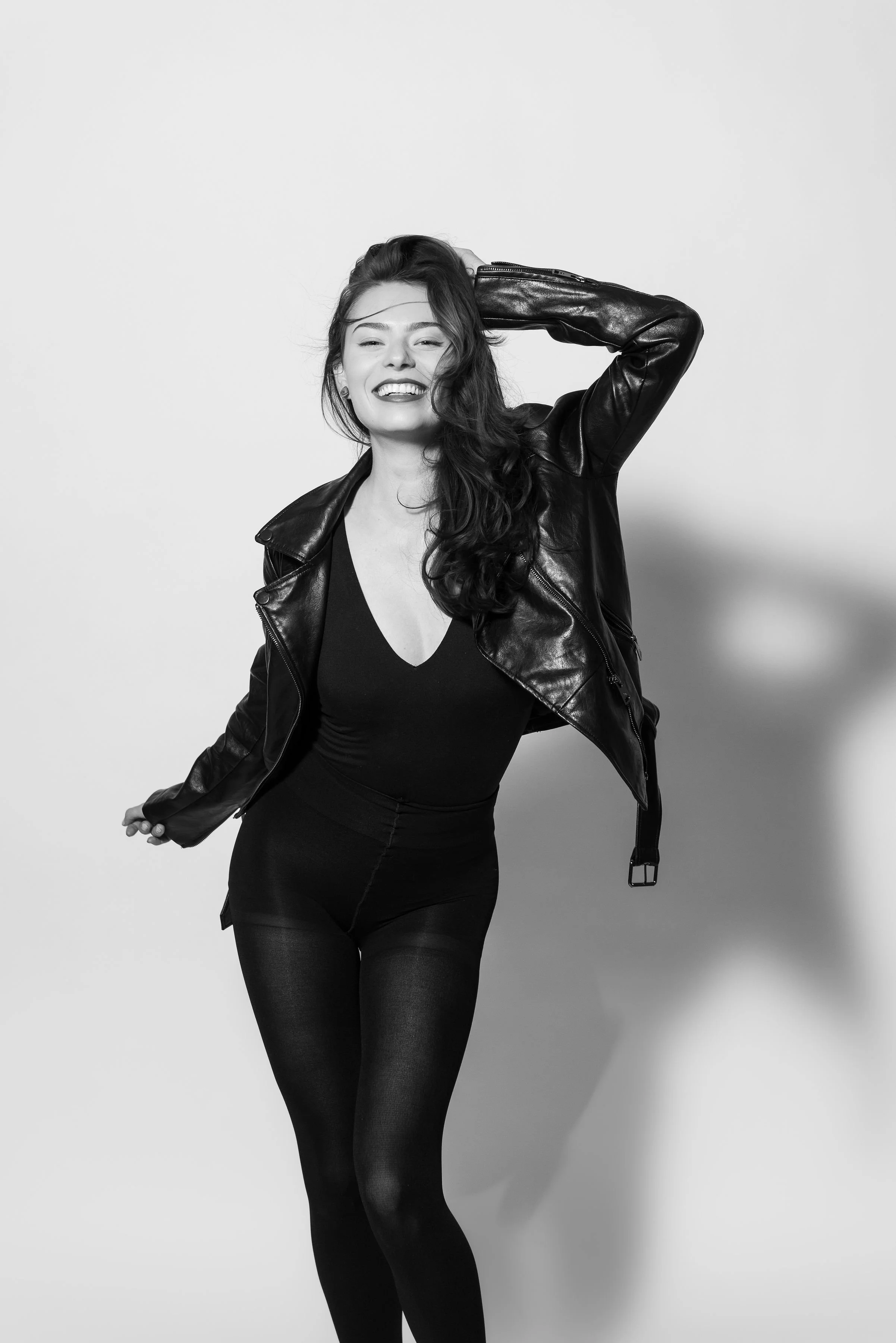 A woman with long wavy hair wearing a leather jacket, black top, and black tights, smiling and posing with one hand behind her head against a plain background.