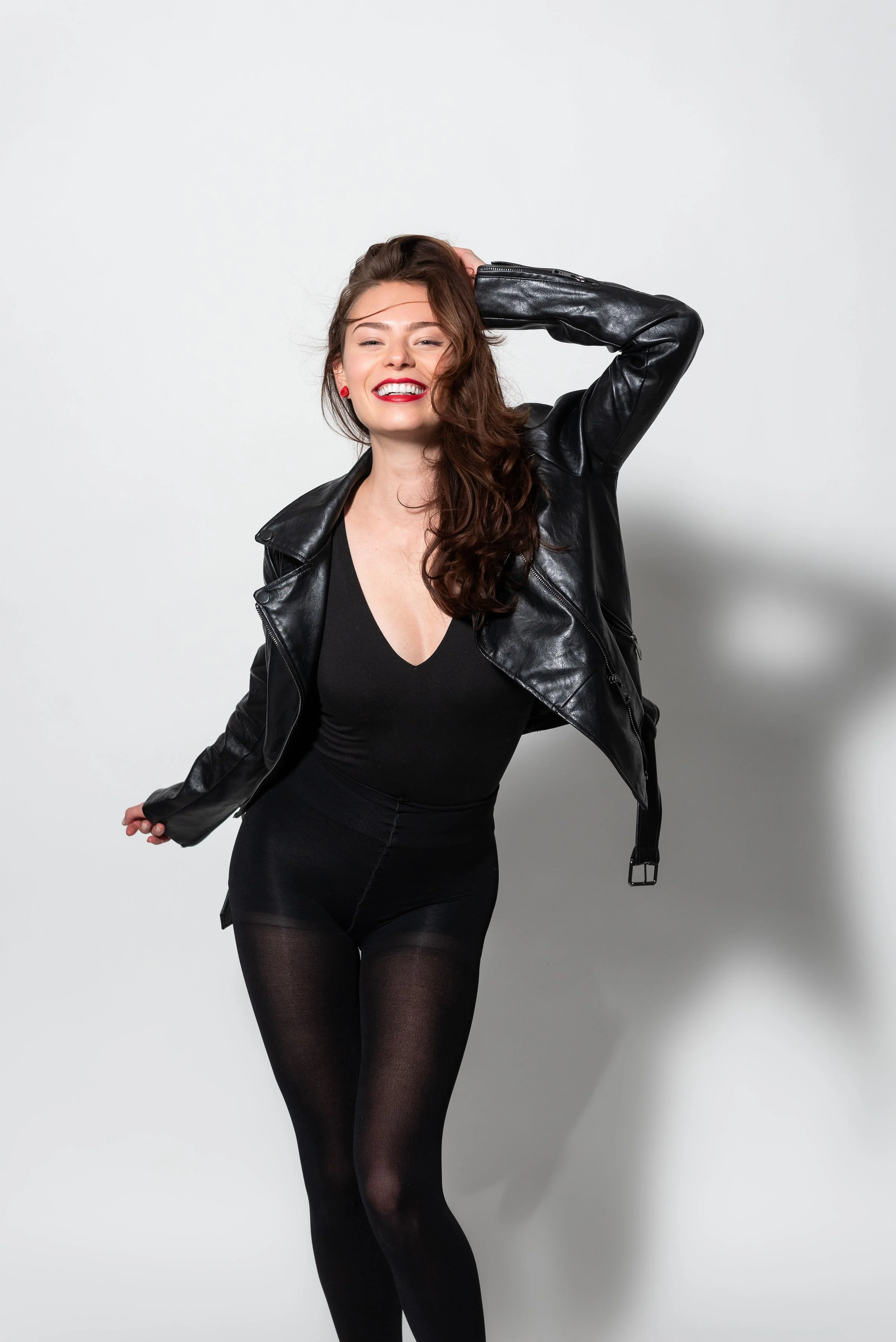 Young woman in black leather jacket, black dress, and sheer black tights, smiling with hand behind her head, standing against a plain light gray background.