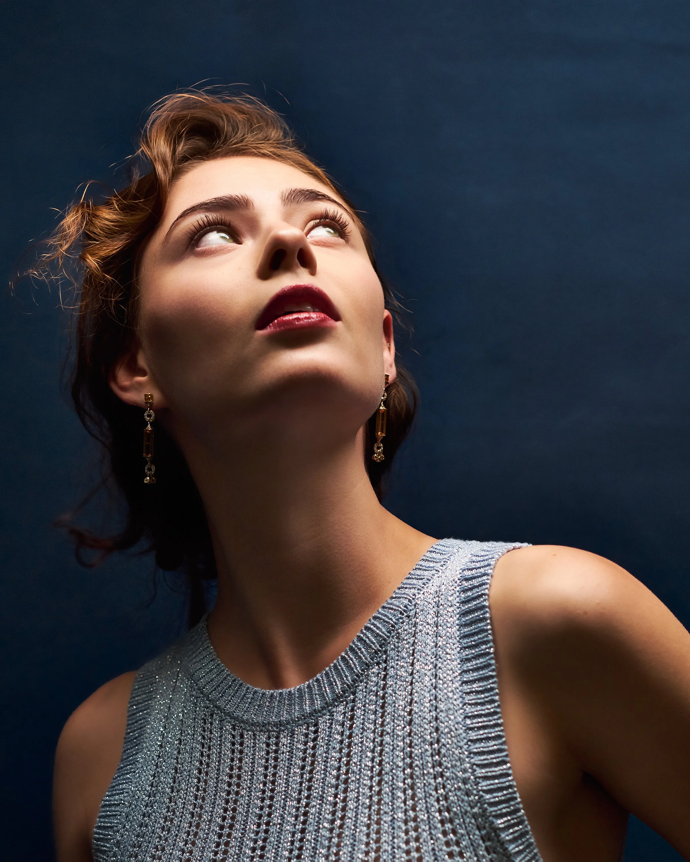 A woman with short brown hair and earrings looking upwards, wearing a silver sleeveless top, against a dark blue background.