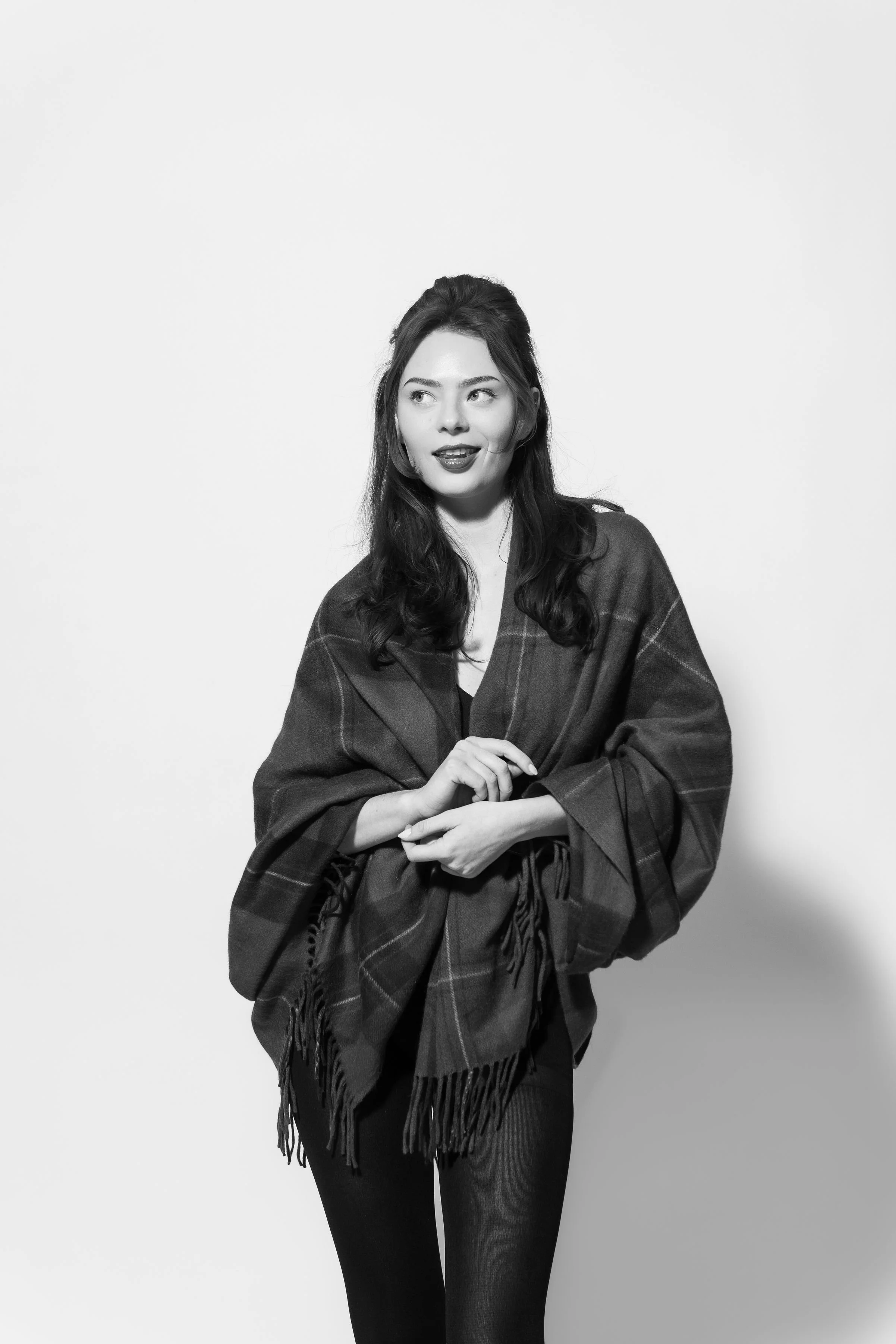 Black and white photo of a woman with long dark hair, wearing a plaid shawl, dark pants, standing against a plain light background, looking to the side with a slight smile.
