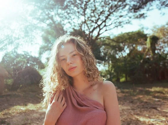 Young woman with curly hair outdoors in sunlight, wearing a pink strapless top, with trees and a bright sky in the background.