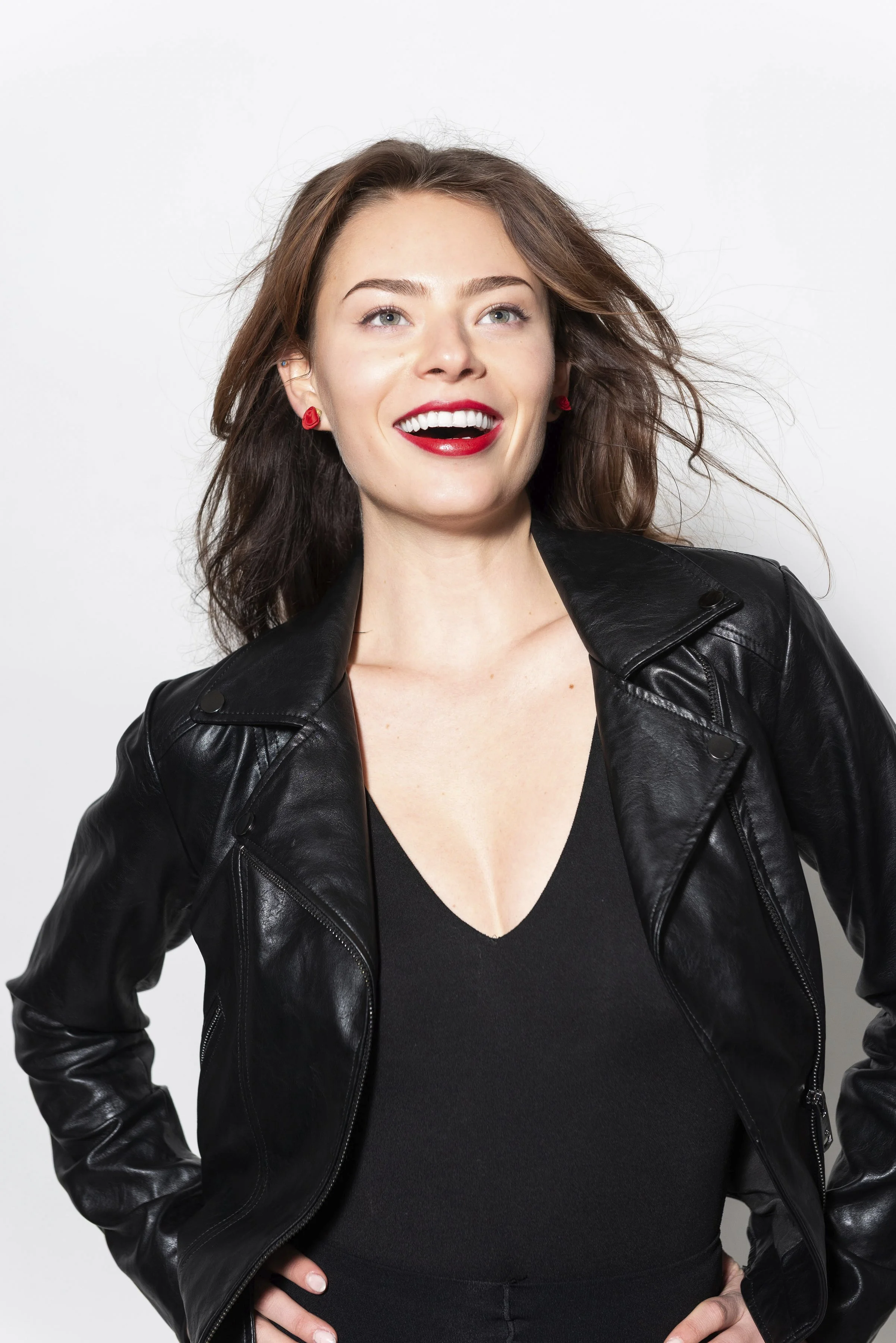 A woman wearing a black leather jacket and red lipstick, smiling with her hand on her hip, against a plain white background.