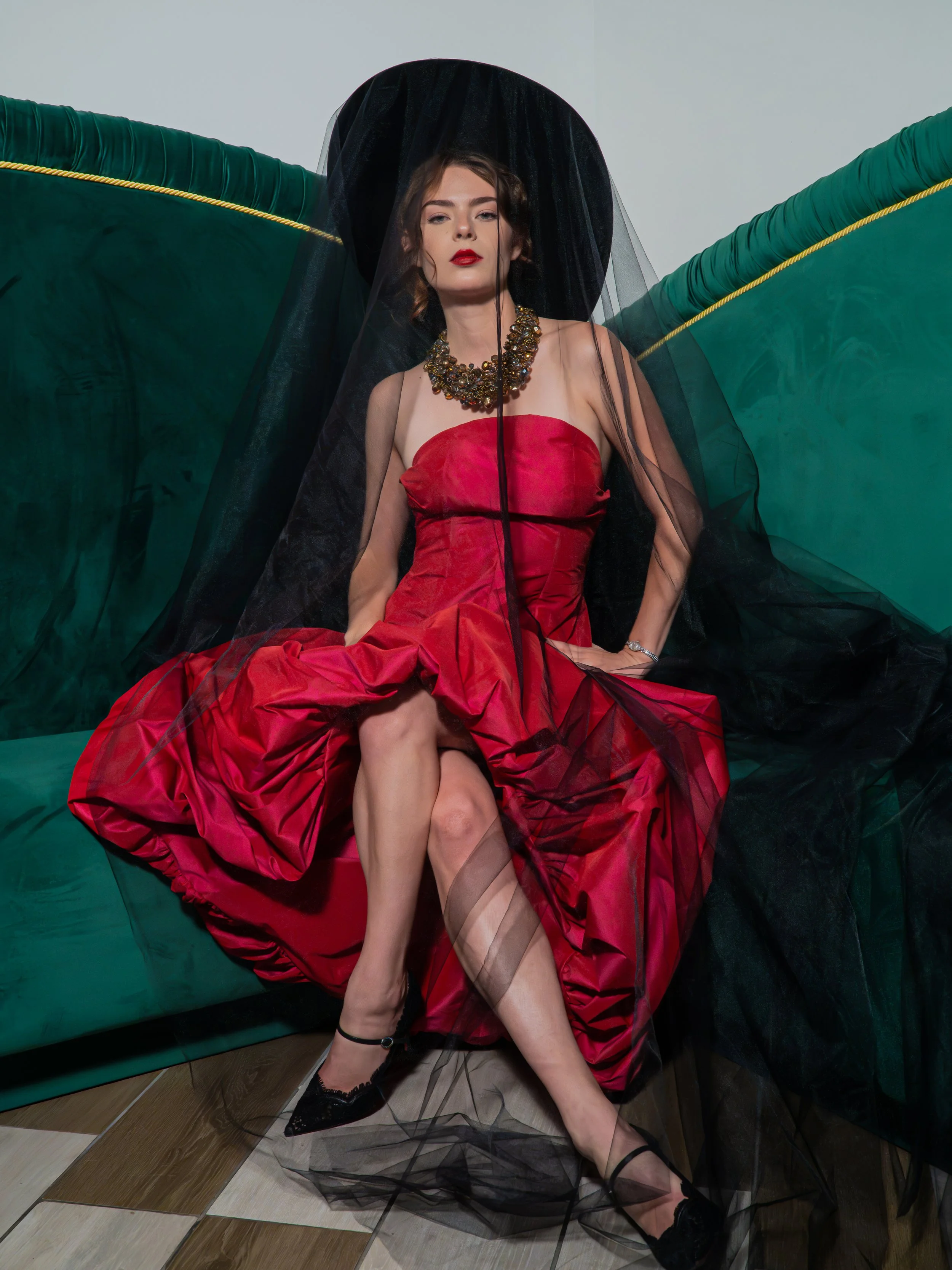 A woman dressed in a red gown, wearing a black hat with black veil, and a large statement necklace, sitting on a green sofa with black tulle fabric around her, with her legs crossed, looking directly at the camera.