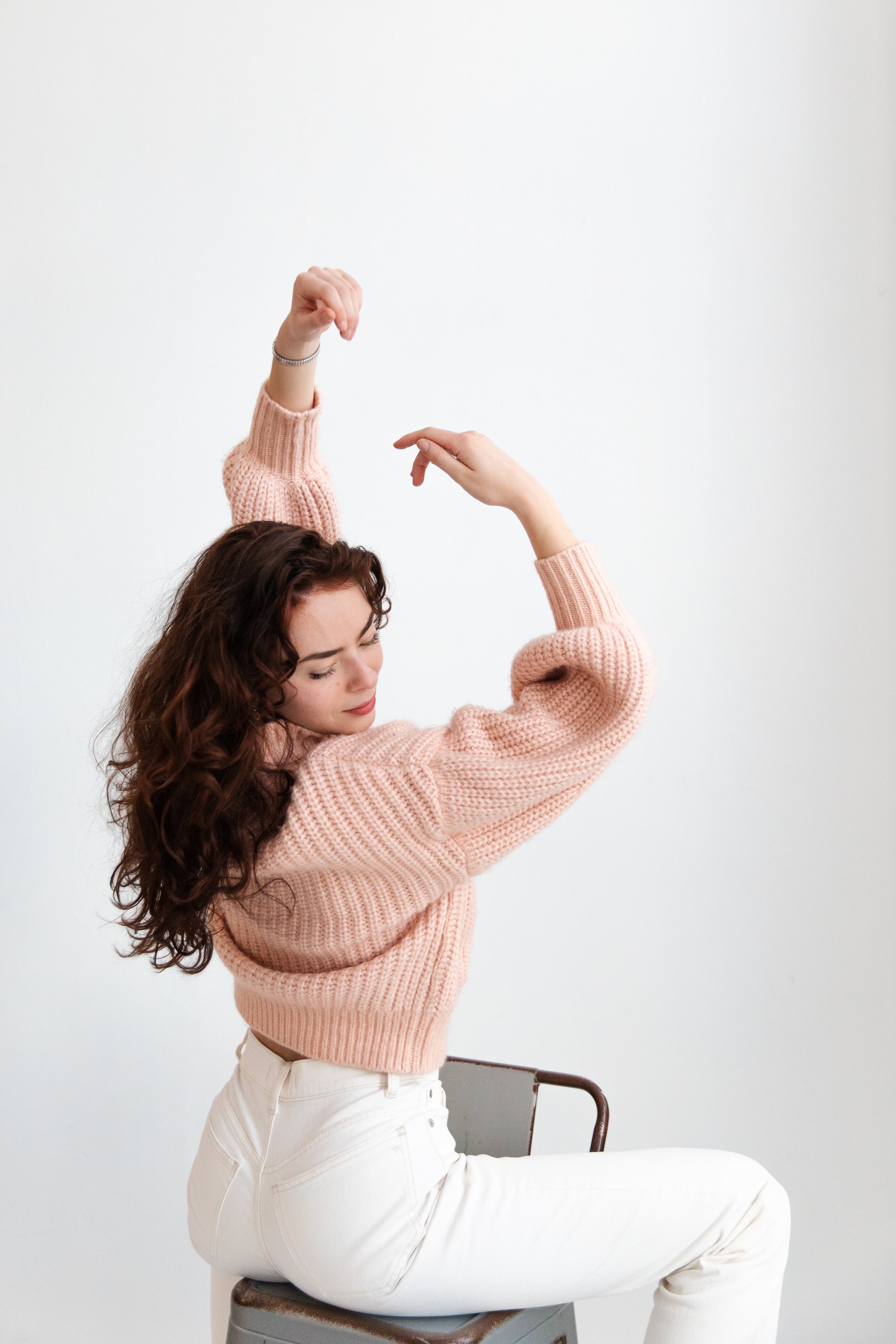 A woman with curly brown hair is sitting on a chair, wearing white pants and a pink knitted sweater. She is posing with her left arm bent and raised, and her right arm curved downward, with her eyes closed.