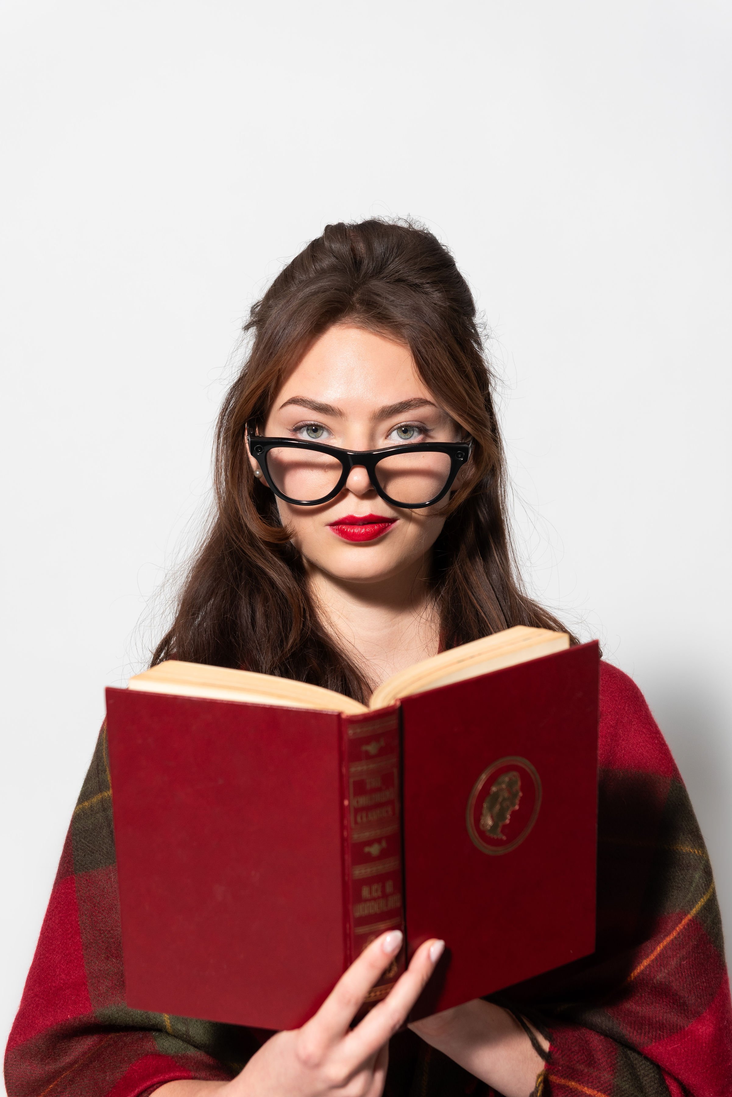 A young woman with brown hair and blue eyes wearing large black glasses and red lipstick is reading a red hardcover book.