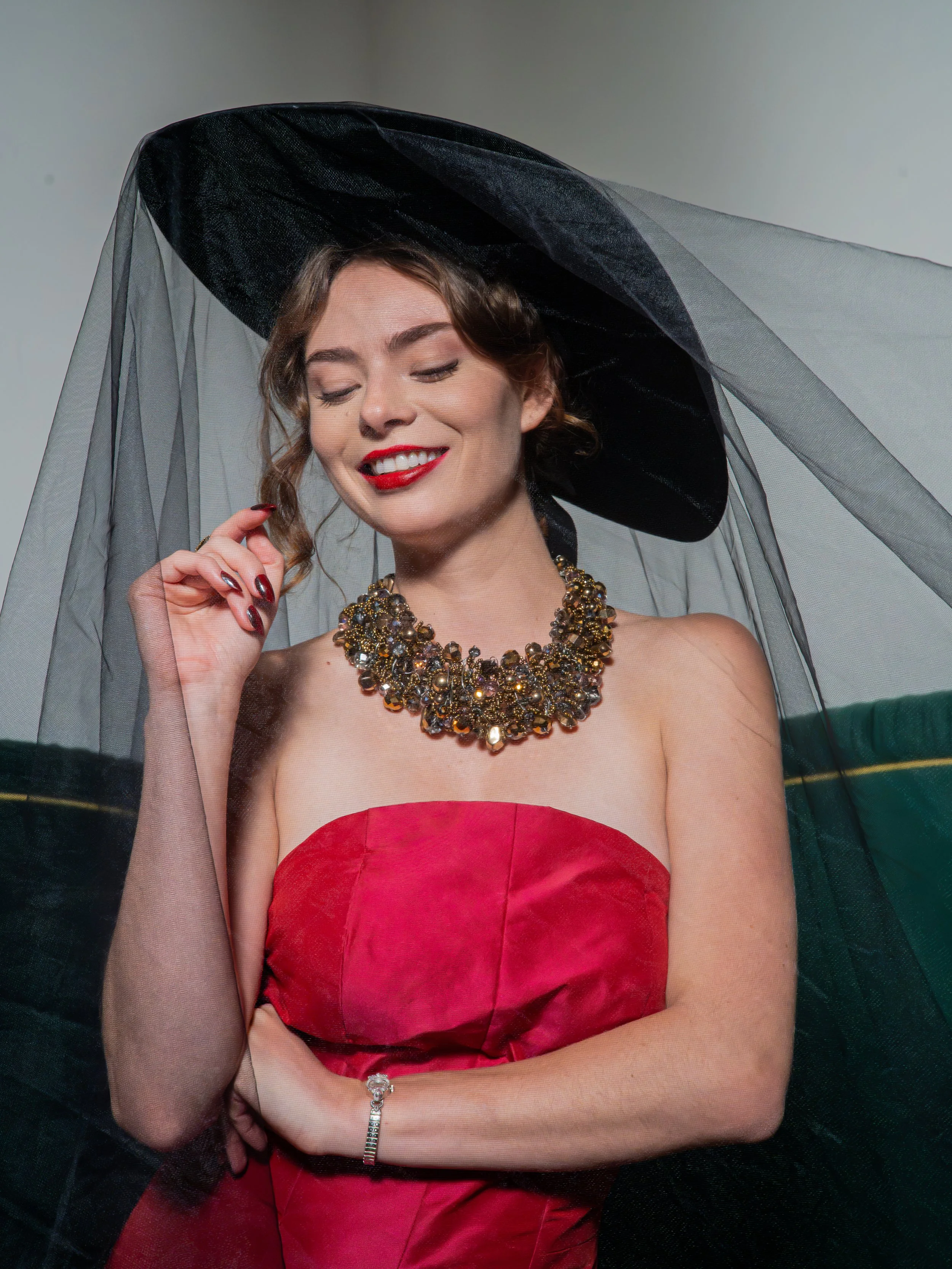 A woman with makeup and curly hair, wearing a large black hat with a veil, a red satin strapless dress, and a multi-layered beaded necklace, standing in front of a plain background.