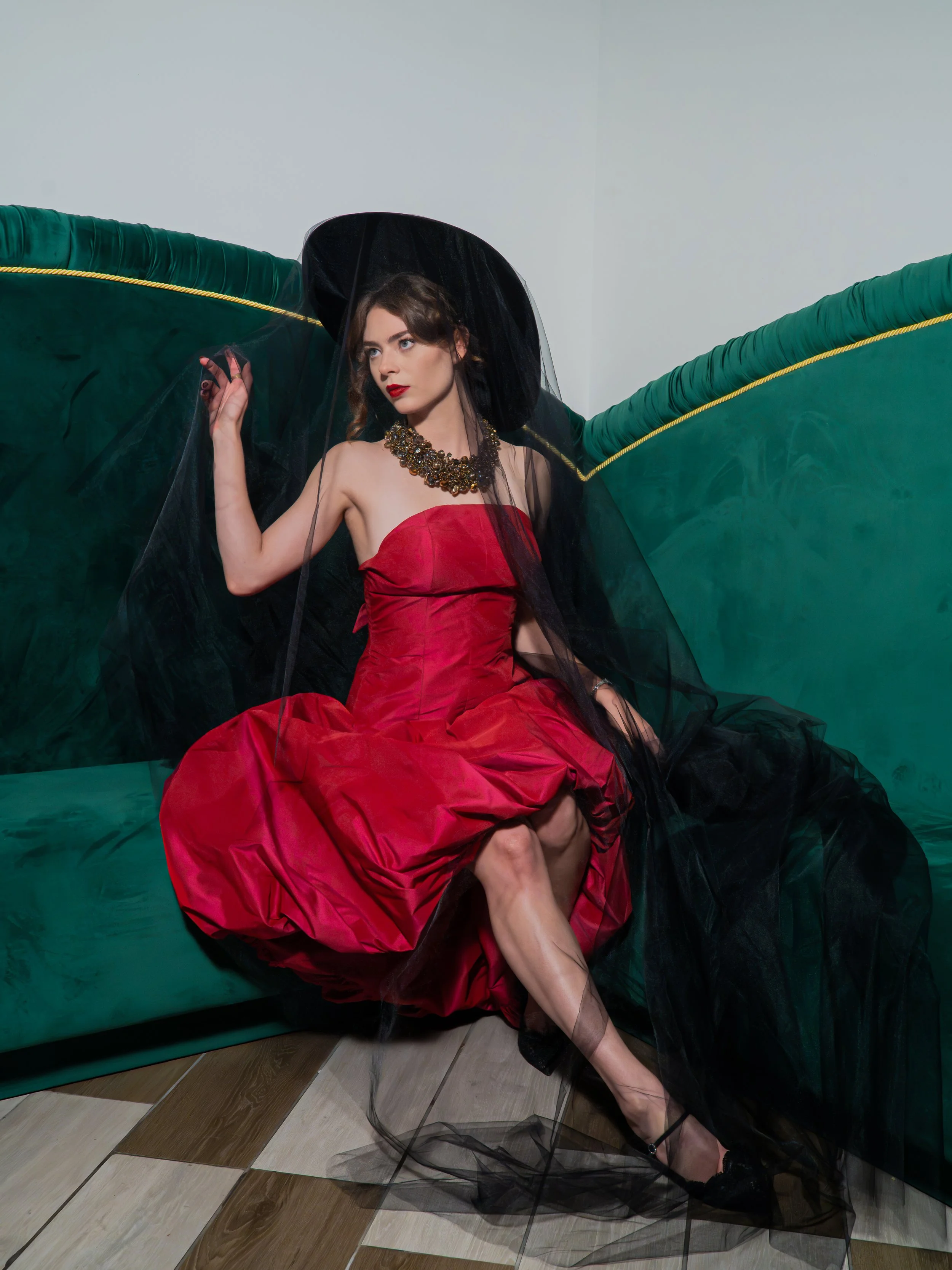 A woman in a strapless red gown with black tulle and a statement necklace sits on the floor against a green velvet seating with gold trim, with her legs crossed and one arm raised. She is wearing black high heels and has a dramatic makeup look with b
