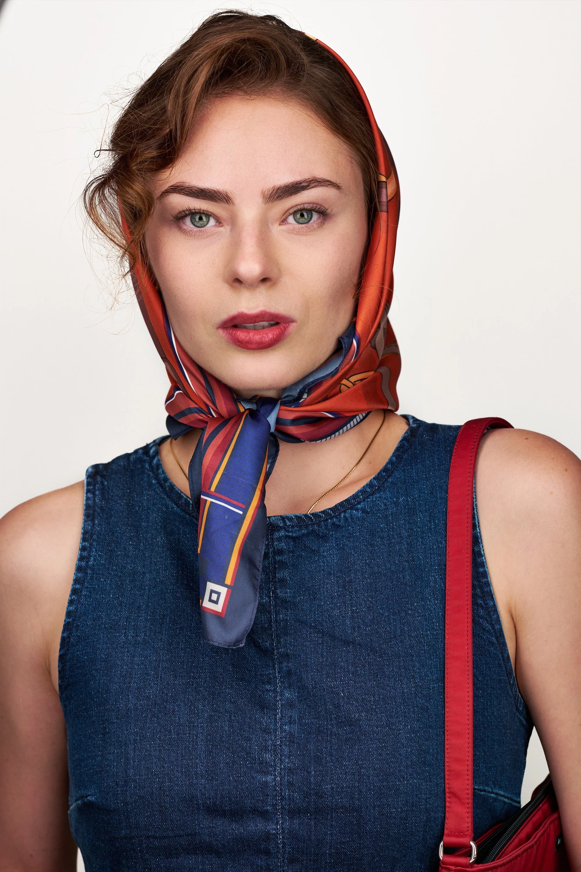 A woman with curly brown hair, green eyes, and fair skin wearing a red and blue headscarf, a sleeveless denim top, and carrying a red bag.