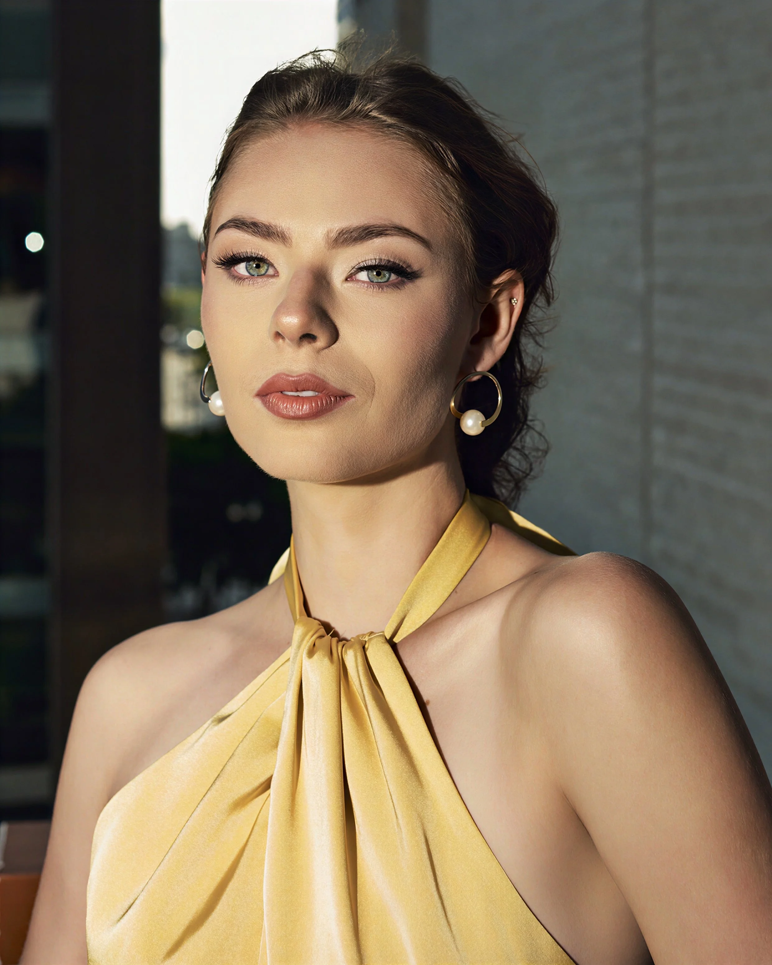 Close-up of a woman with short brown hair, green eyes, and wearing pearl earrings and a yellow halter dress, standing outdoors near a building with a gray tiled wall.