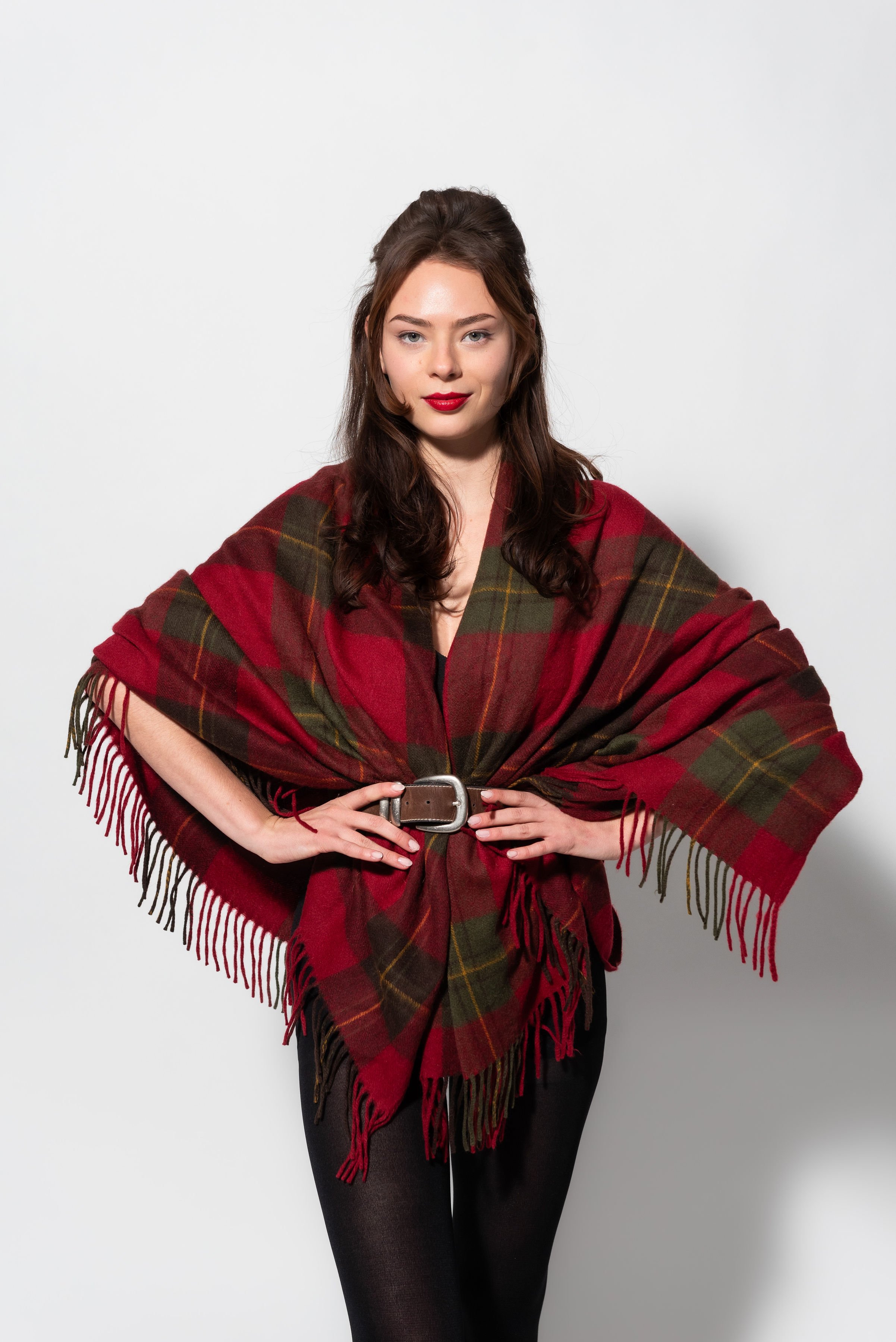 A woman with long dark hair in loose waves, wearing red lipstick, a dark top, black pants, and a plaid shawl with red, green, and brown colors and fringe, standing against a plain white background.
