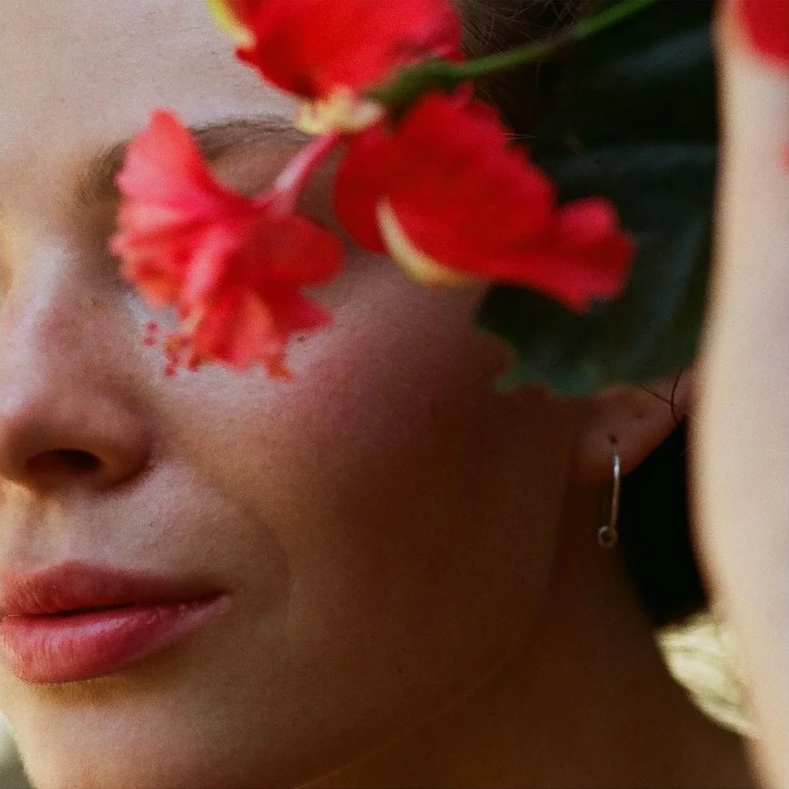 Close-up of a woman with red flowers obscuring part of her face, including her lips and one eye, wearing a small hoop earring.