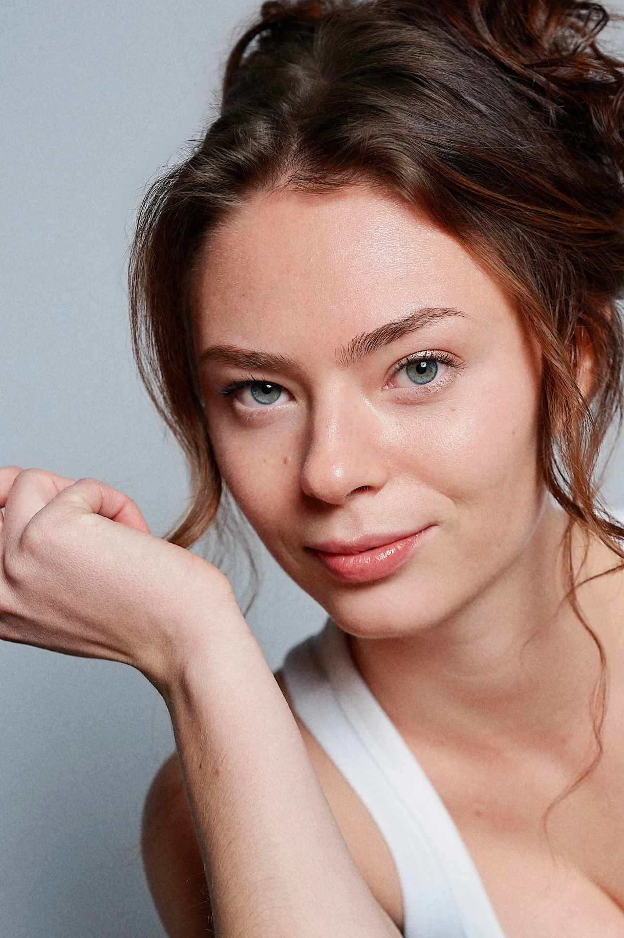 Close-up of a young woman with short, wavy, auburn hair and blue eyes, smiling gently at the camera, with her left hand resting near her face, against a gray background.