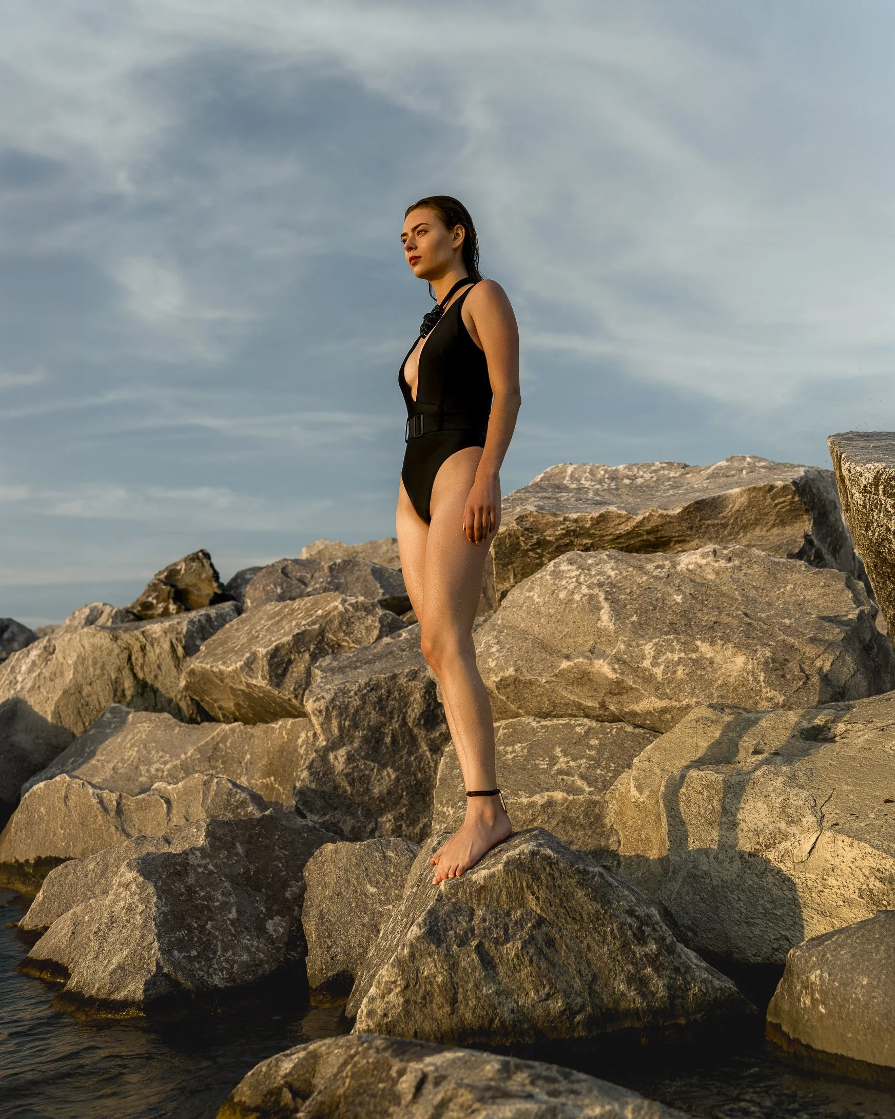 A woman in a black swimsuit standing on large rocks by the water during sunset or sunrise.