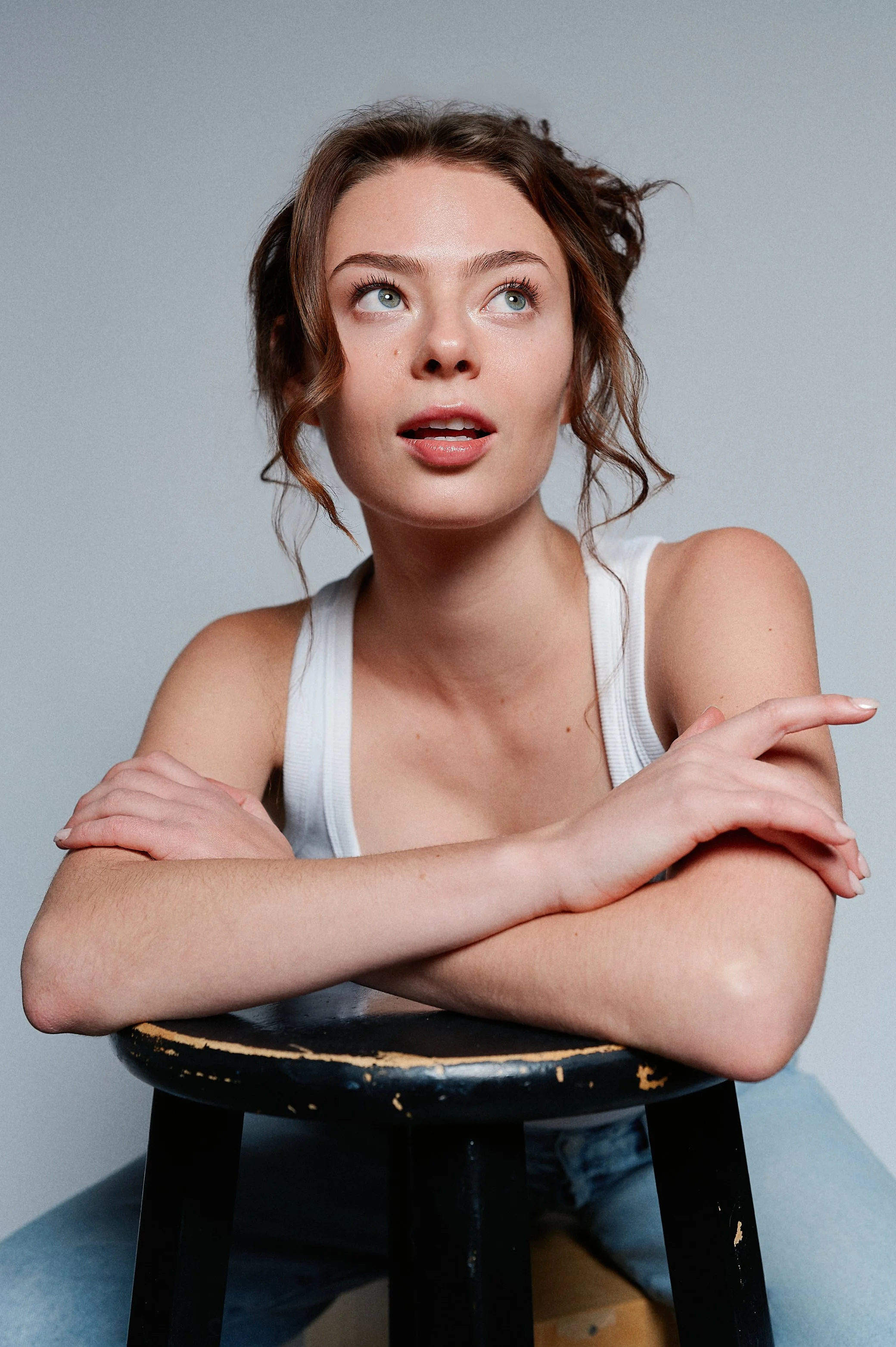 A woman with wavy brown hair and green eyes looking to her right, wearing a white tank top, sitting on a black wooden stool, arms crossed on a black table in front of her, against a plain gray background.
