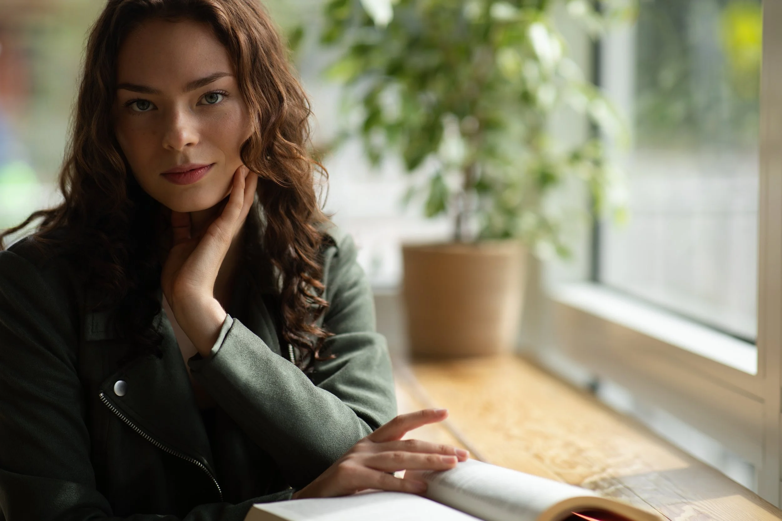 A young woman with curly brown hair looking at the camera while sitting at a wooden table near a large window with greenery outside, resting her chin on her hand.